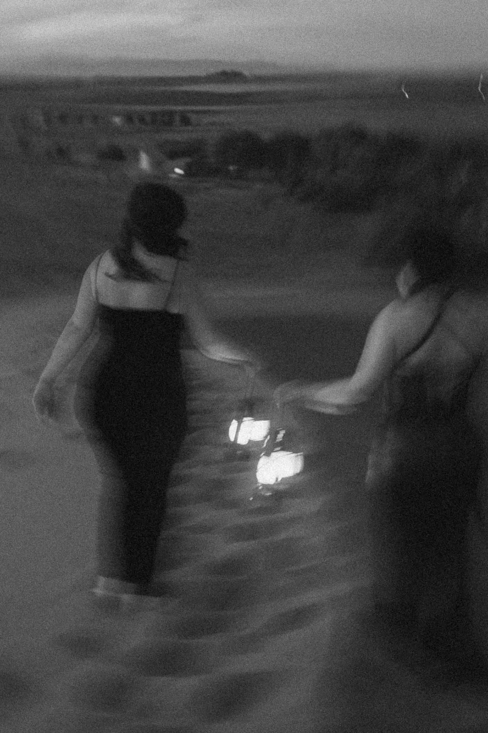  Two women walk side by side in the dark, each holding a lantern. The image is black and white, softly blurred, and appears to be taken outdoors on a sandy path with distant lights and landscape in the background. 