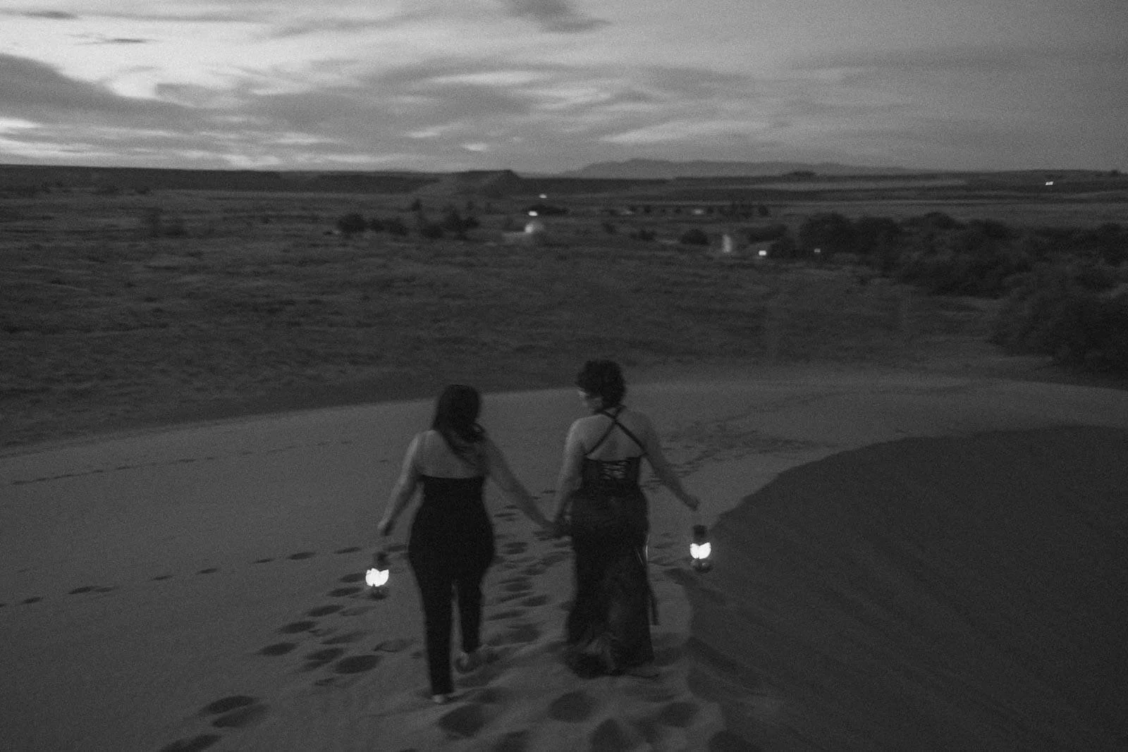  Two women in black dresses hold hands and lanterns, walking up a sandy hill at dusk. Their backs are to the camera and footprints trail behind them. The landscape is open with distant lights and cloudy skies. 