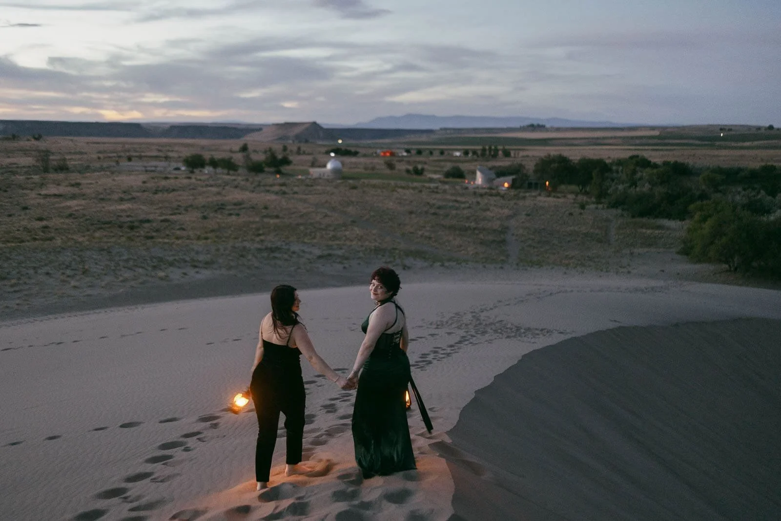  Two women in dark clothing hold hands and lanterns, standing on sand dunes at dusk, looking back toward the camera. A vast landscape with scattered lights and distant buildings stretches out behind them. 