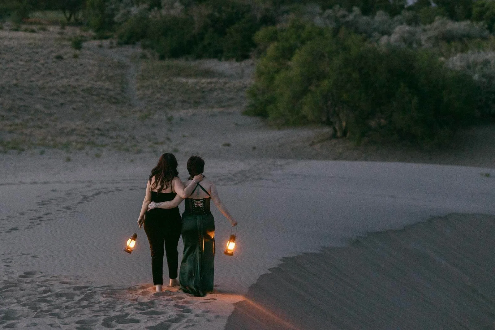  Two women walk side by side on sandy dunes at dusk, each holding a lantern. They have their arms around each other, surrounded by sparse vegetation and distant trees. 