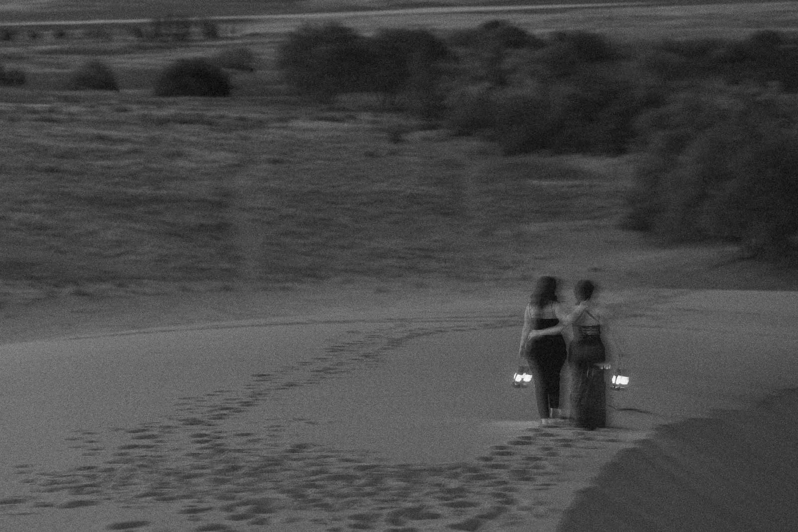 Two women walk away on a sandy path at dusk, each holding a lantern. Their footprints trail behind them, and trees and fields are visible in the background. The image is in black and white. 