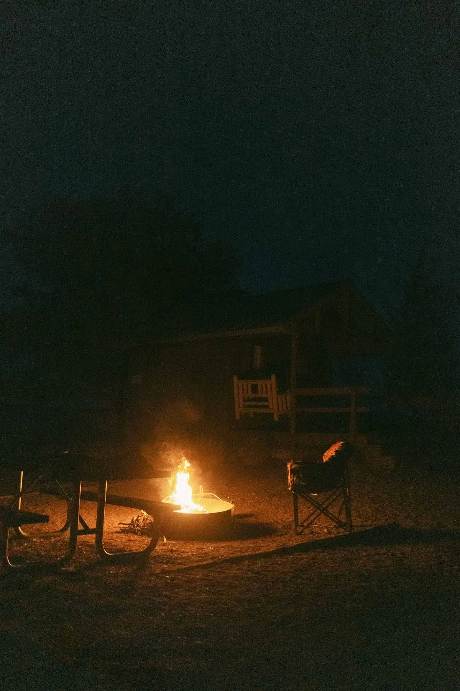  A campfire burns in a fire pit at night, with an empty camping chair and picnic table nearby. In the background, a dark cabin is visible under the night sky. 