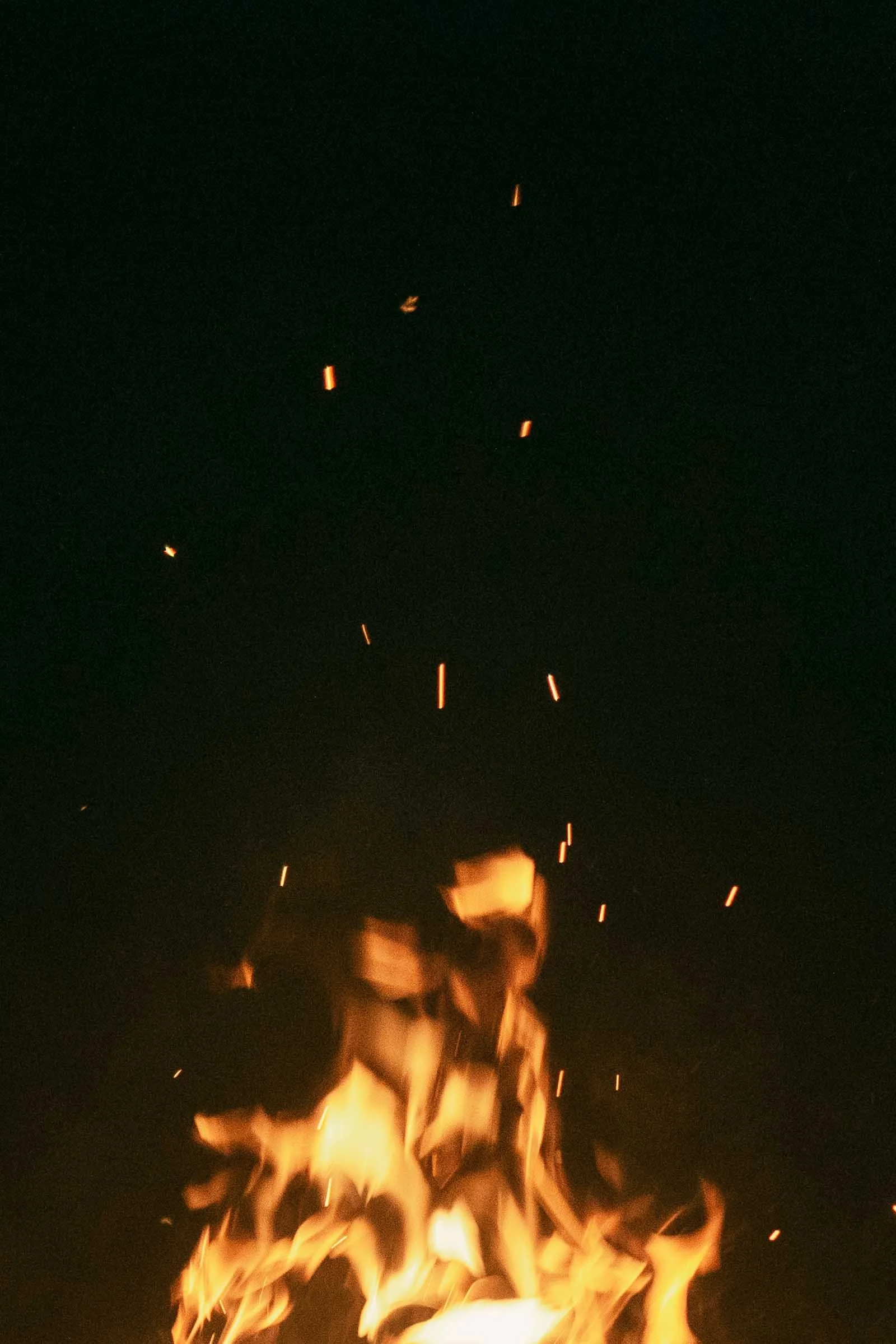  Bright orange flames lick upward through glowing embers against a deep black night sky, a campfire burning strong during an overnight engagement session at Bruneau Sand Dunes. 