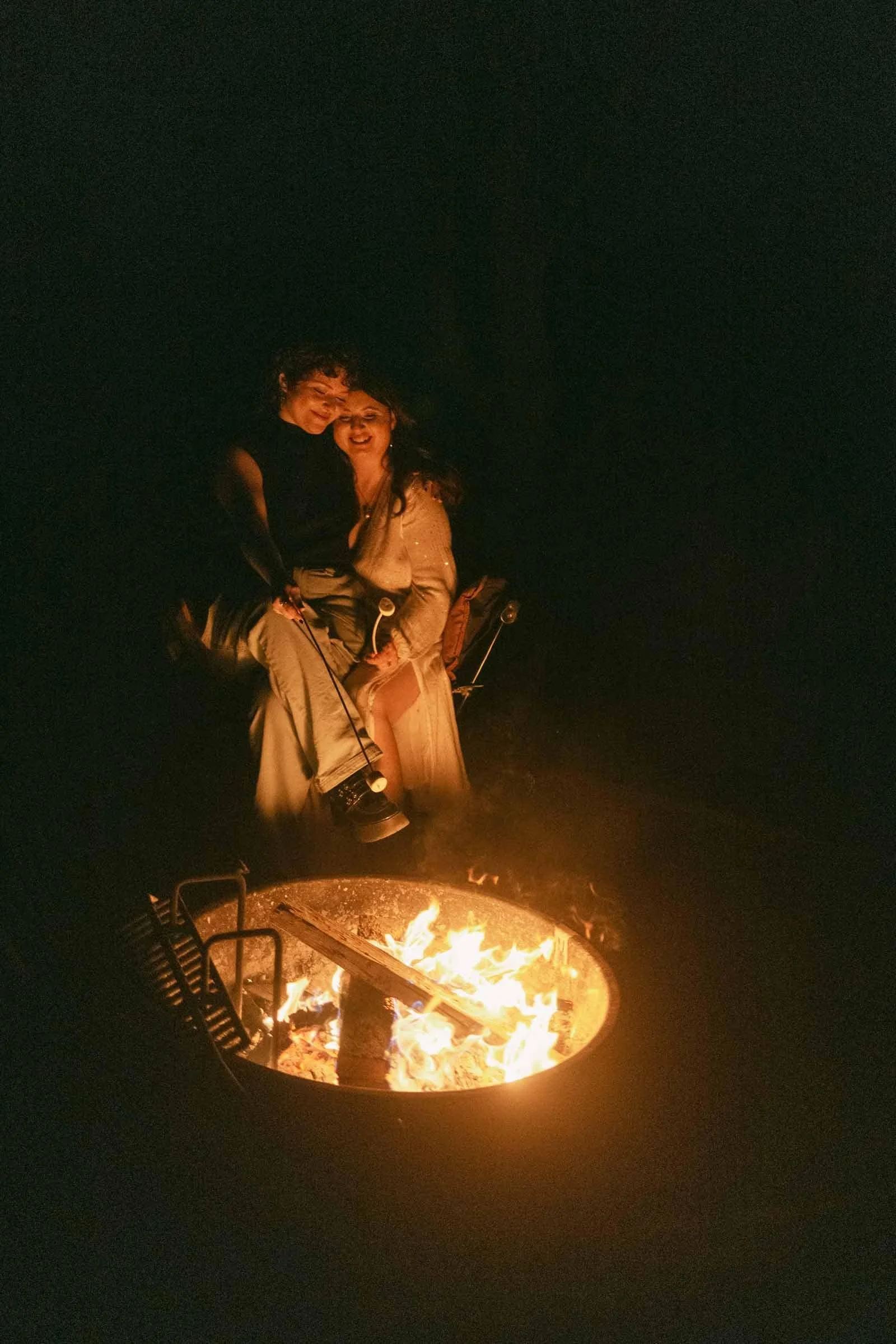  Two women sit close together by a campfire at night, illuminated by the fire’s warm glow. One woman sits on the other’s lap, both smiling and looking happy and relaxed outdoors. 