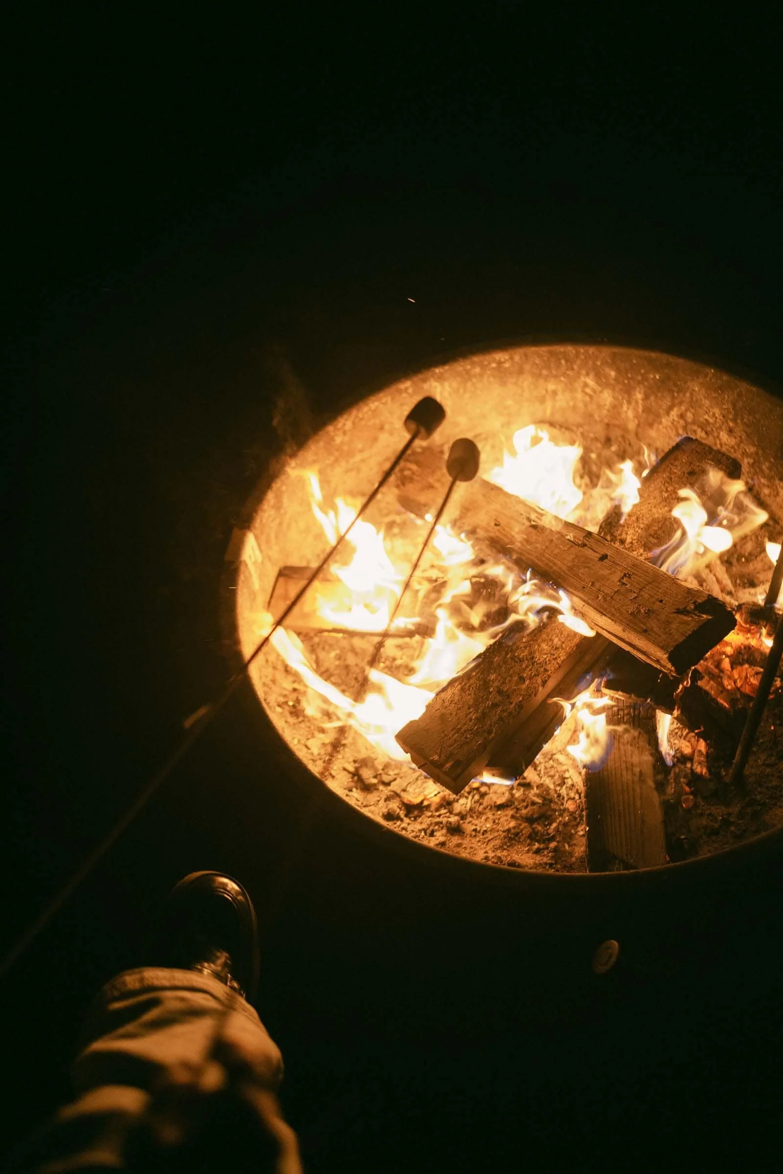  Two marshmallows on sticks are roasting over a campfire with logs burning inside a fire ring at night; a person’s foot is visible in the bottom left corner. 