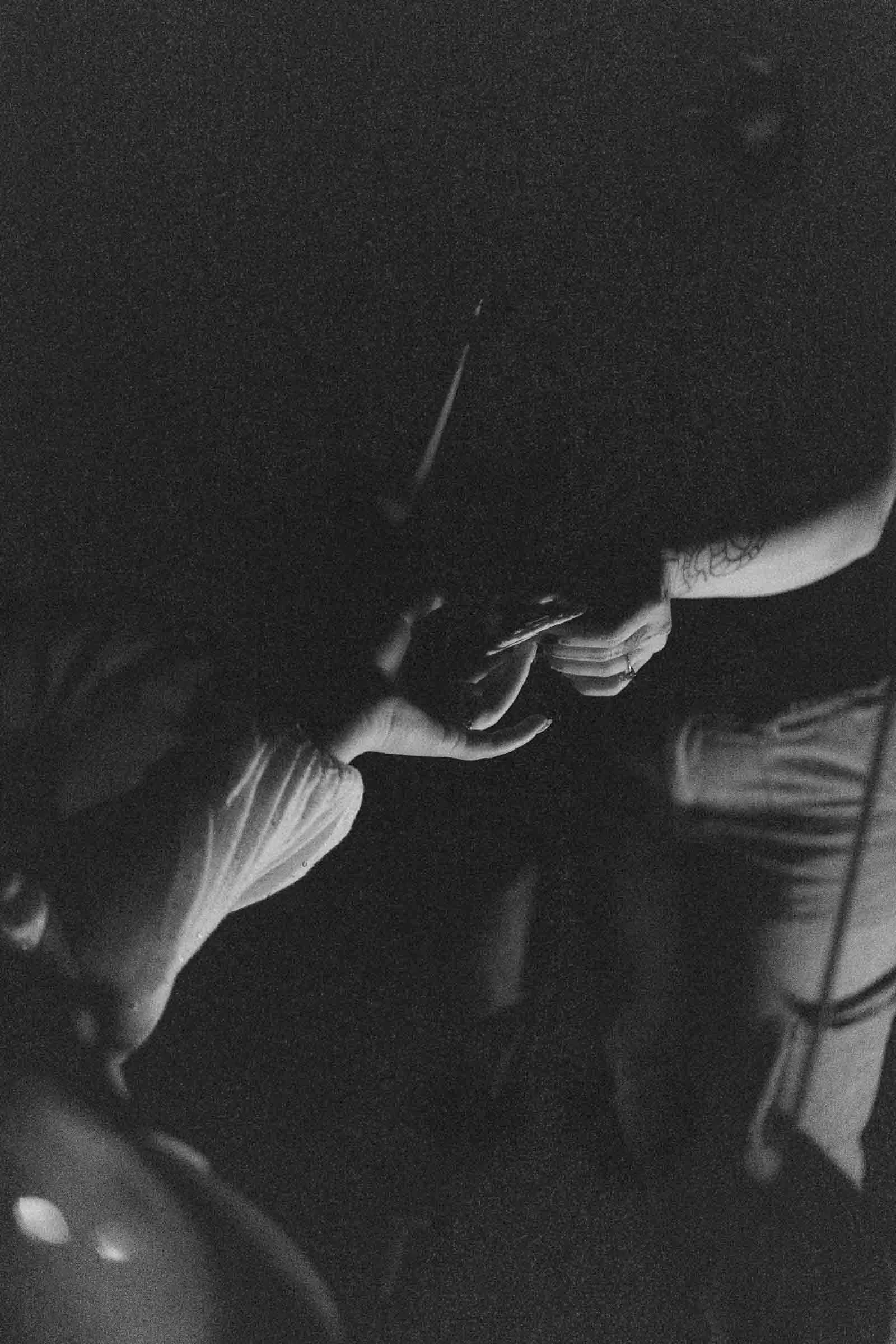  A black-and-white photo shows two people’s hands meeting in the dark, one holding a s’more. Only arms and hands are visible, with tattoos on one arm, creating a moody, intimate atmosphere. 
