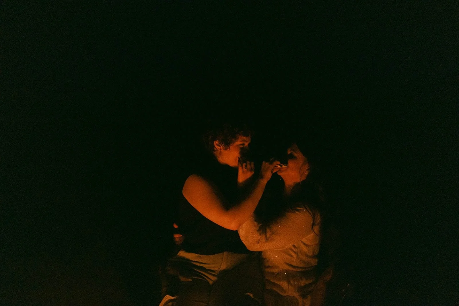  Two women sit closely together in low light, gently touching each other’s faces and looking at each other, with a warm glow illuminating them against a dark background. 