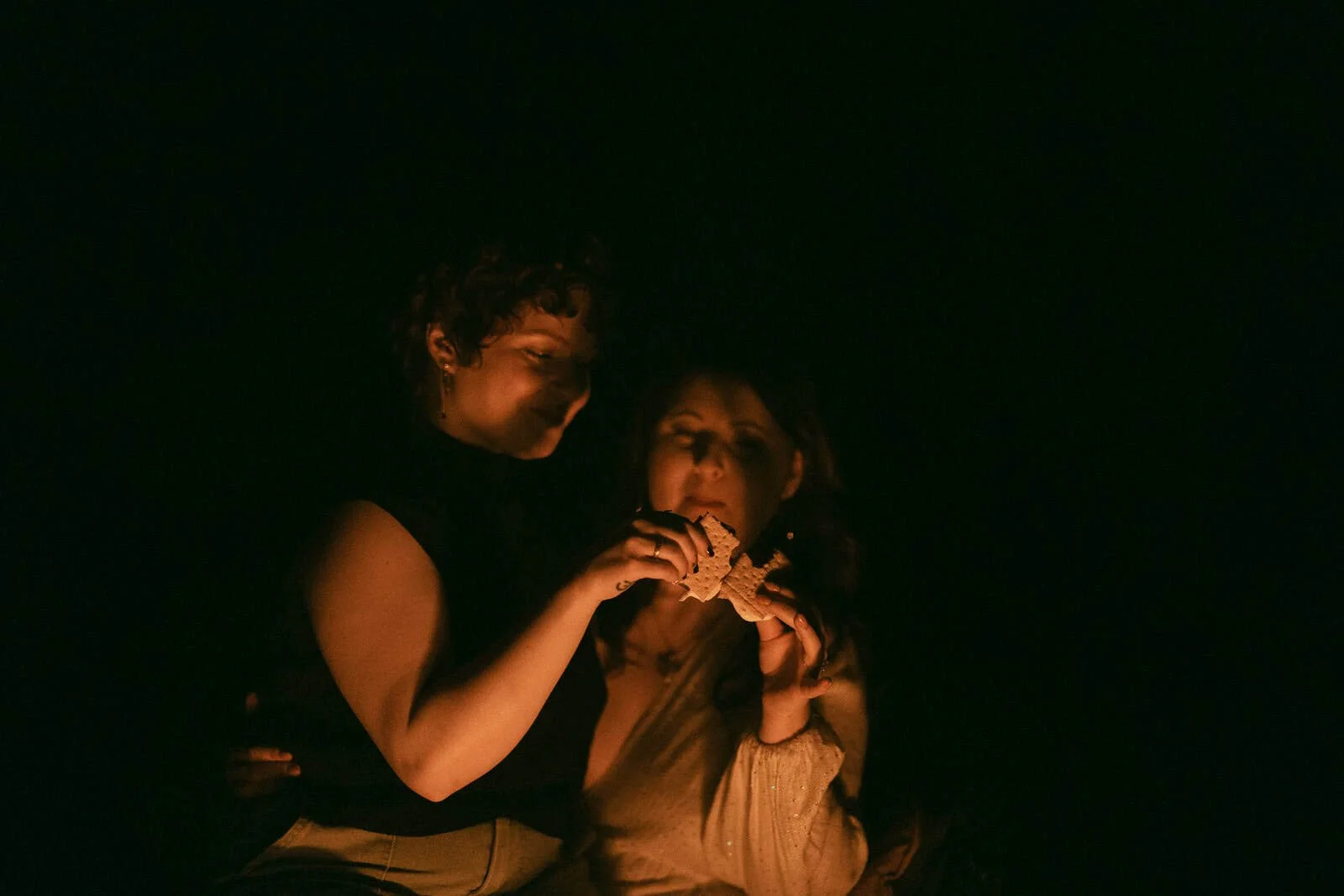  Two women sit close together in dim light, illuminated by a fire, holding a s’more together and gazing at it, their faces partially visible in the glow. 