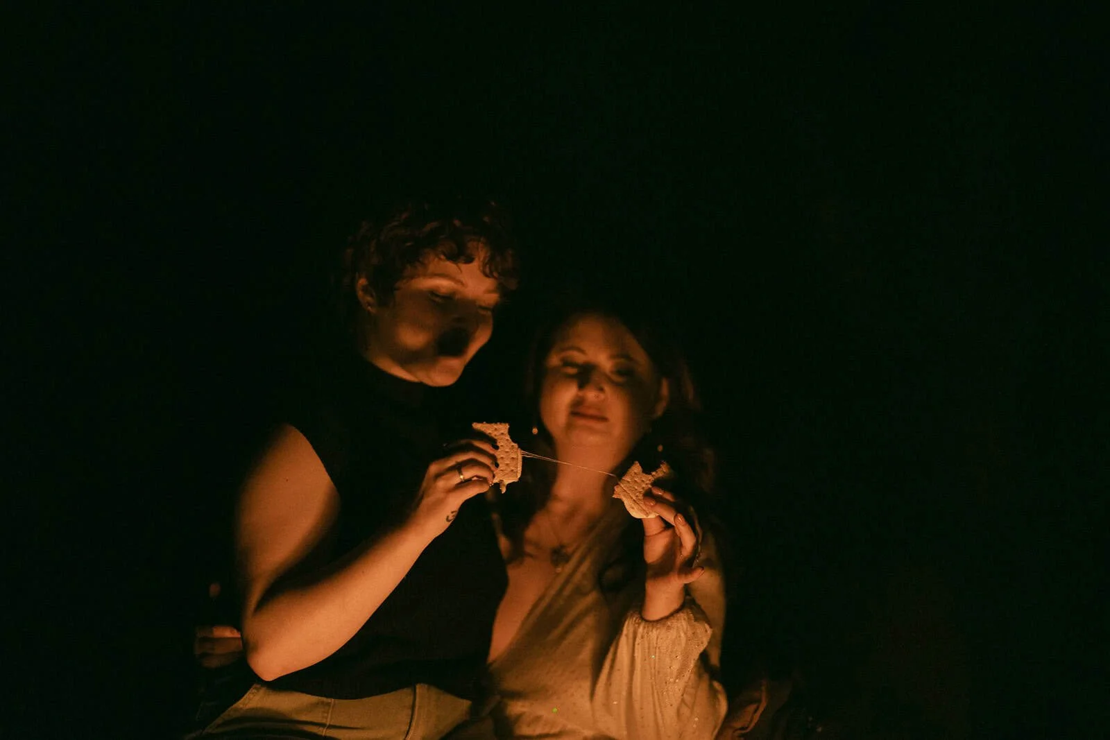  Two people sit close together in dim light, holding marshmallows and toasting s’mores over a campfire, their faces softly illuminated by the glow. 