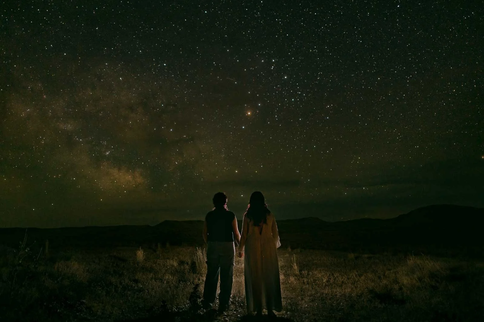 Two women hold hands and gaze upward at a sprawling Milky Way above Bruneau Sand Dunes, Idaho, standing together in the wide open dark of one of the state's best dark sky locations.