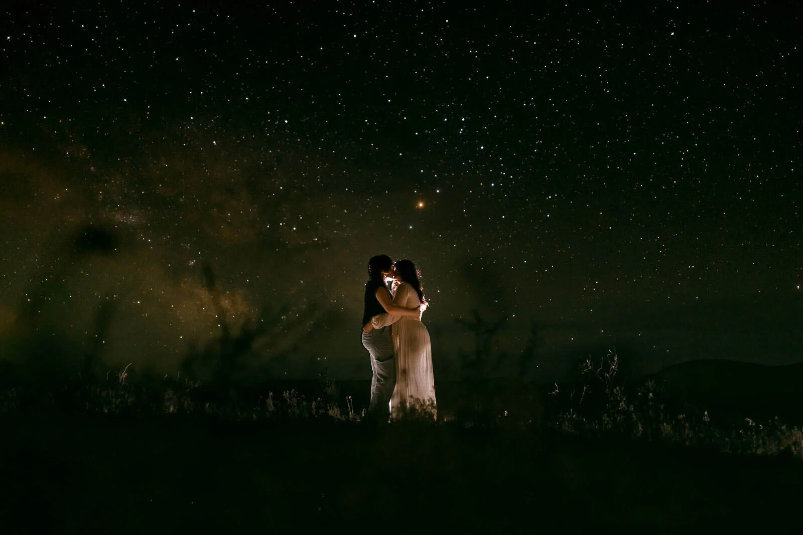  A couple embraces and kisses under a starry night sky, softly illuminated, surrounded by darkness and distant hills. 