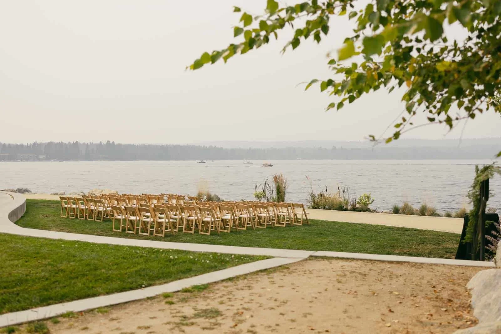 Rows of wooden chairs are set up on a grassy lawn facing a peaceful lake, ready for an outdoor event. Trees and plants border the area under a hazy sky.
