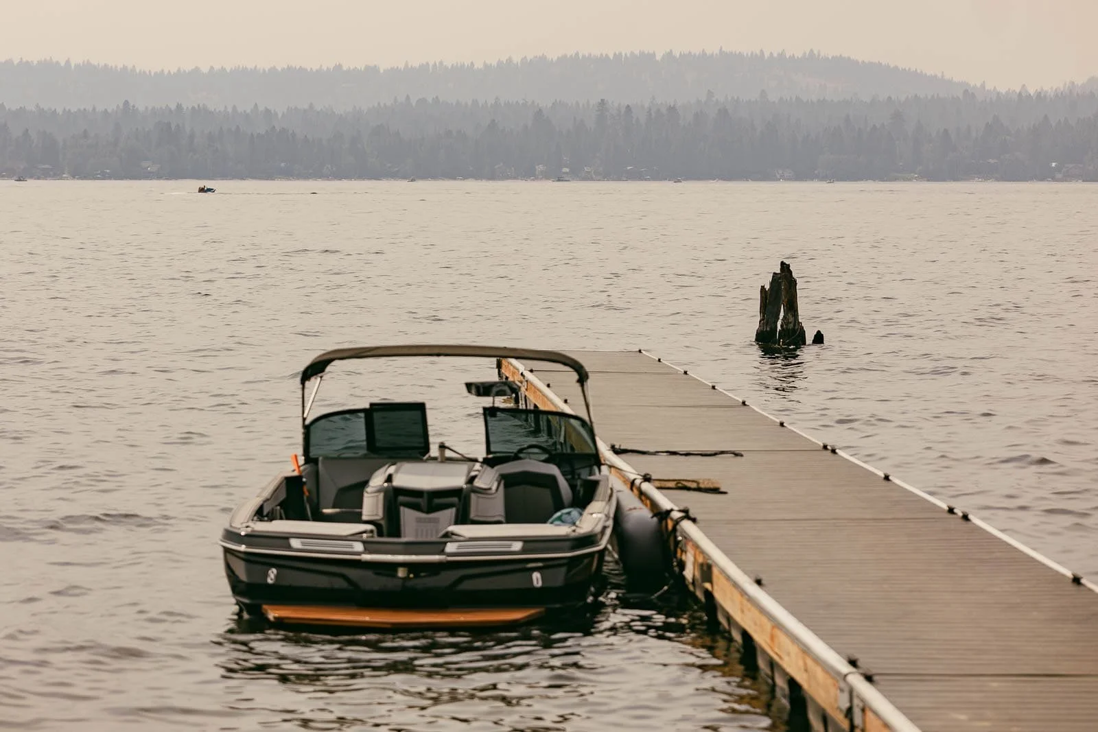 A black motorboat is docked alongside a wooden pier on a calm lake, with forested hills and a hazy sky in the background.