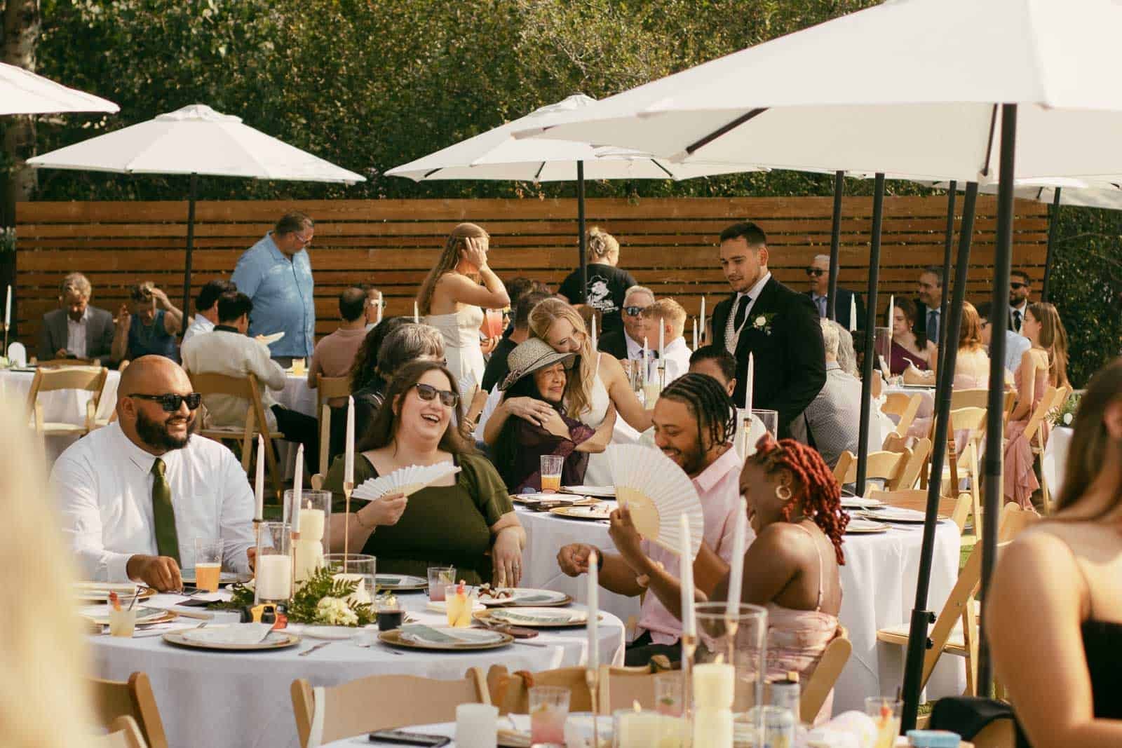 Guests sit at round tables with white tablecloths under outdoor umbrellas, laughing and talking at a sunny daytime event. Some people are holding paper fans. Candles and plates decorate the tables.