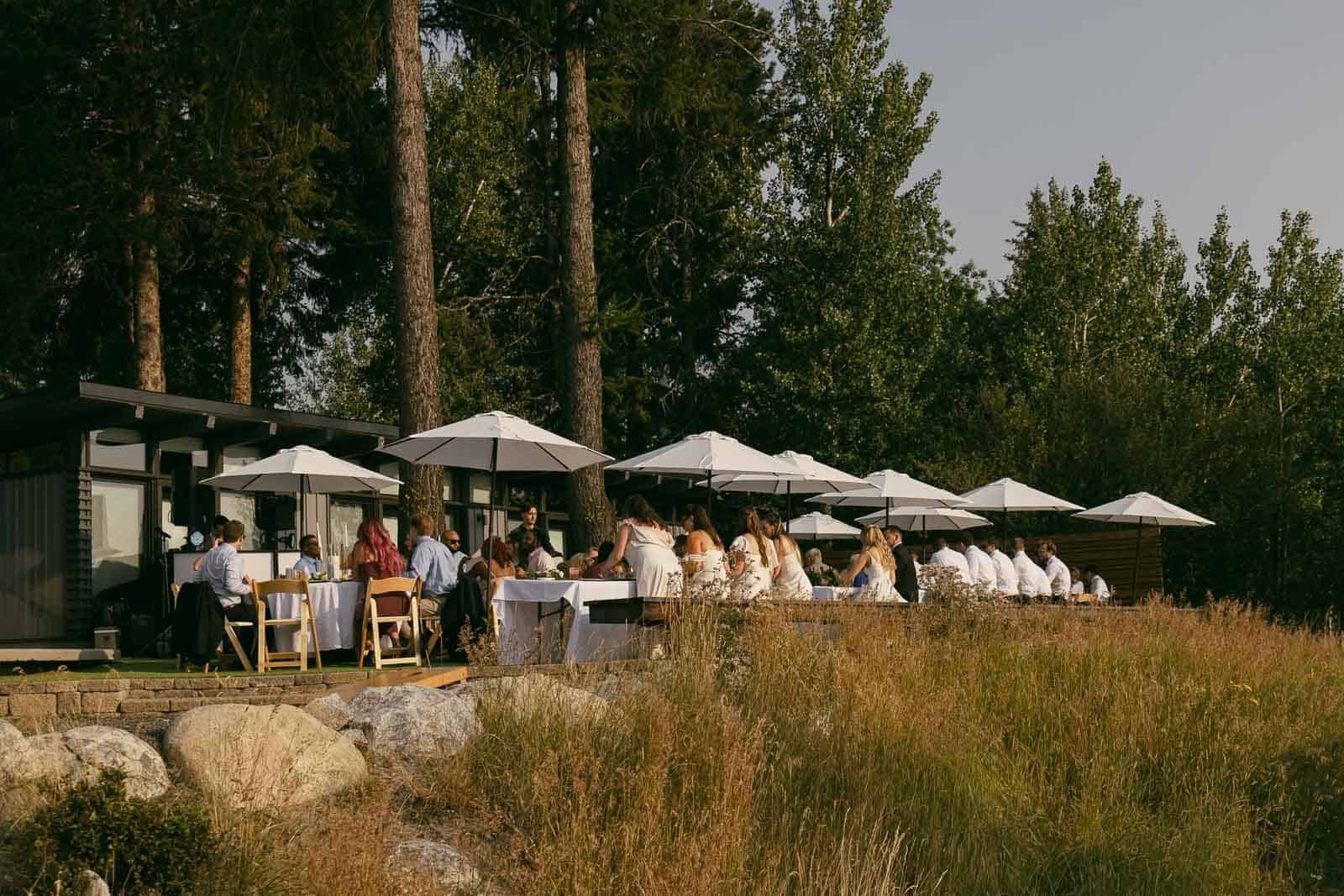 People are dining outdoors at tables with white umbrellas, surrounded by tall trees and grassy landscape, creating a peaceful, natural setting.