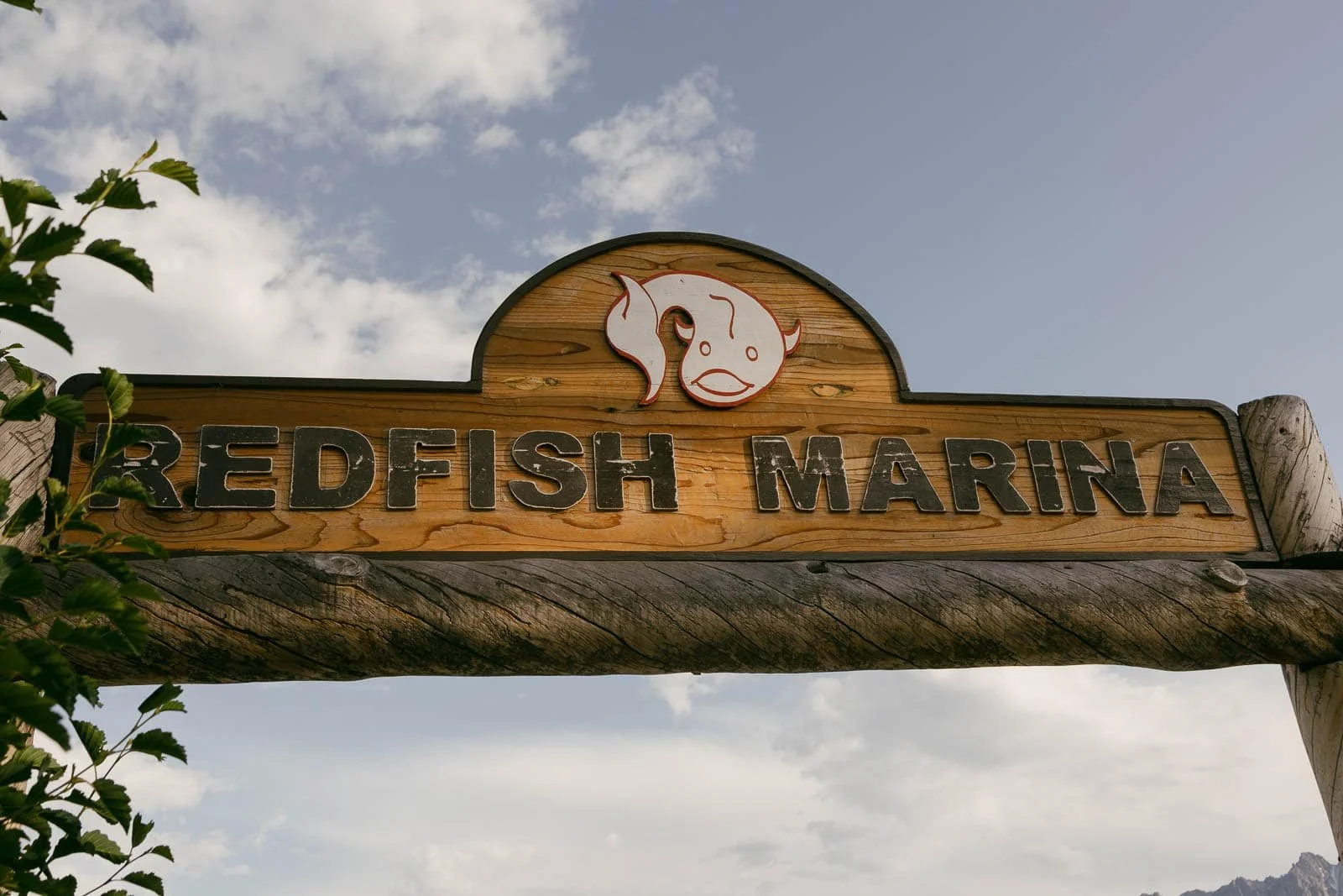  Wooden sign reads “Redfish Marina” with a cartoon fish illustration above the text. Some green leaves are visible on the left, and the sky with clouds is in the background. 