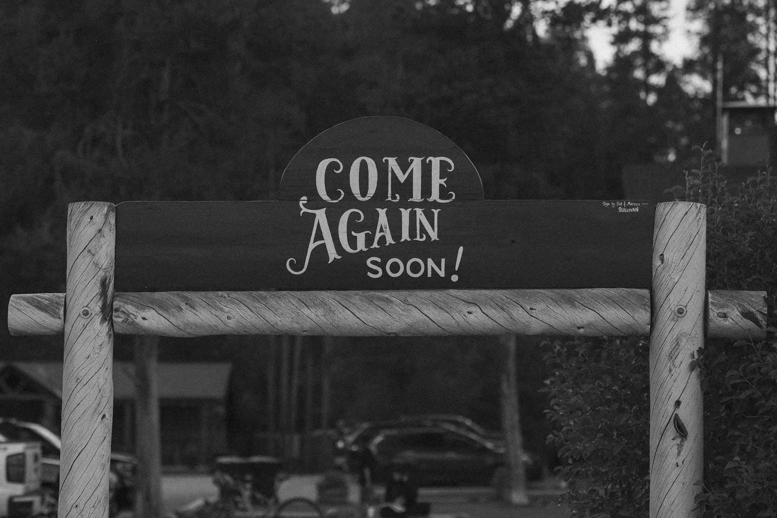  A rustic wooden sign reads “Come Again Soon!” in decorative lettering, set outdoors with trees, parked cars, and a building visible in the background. 