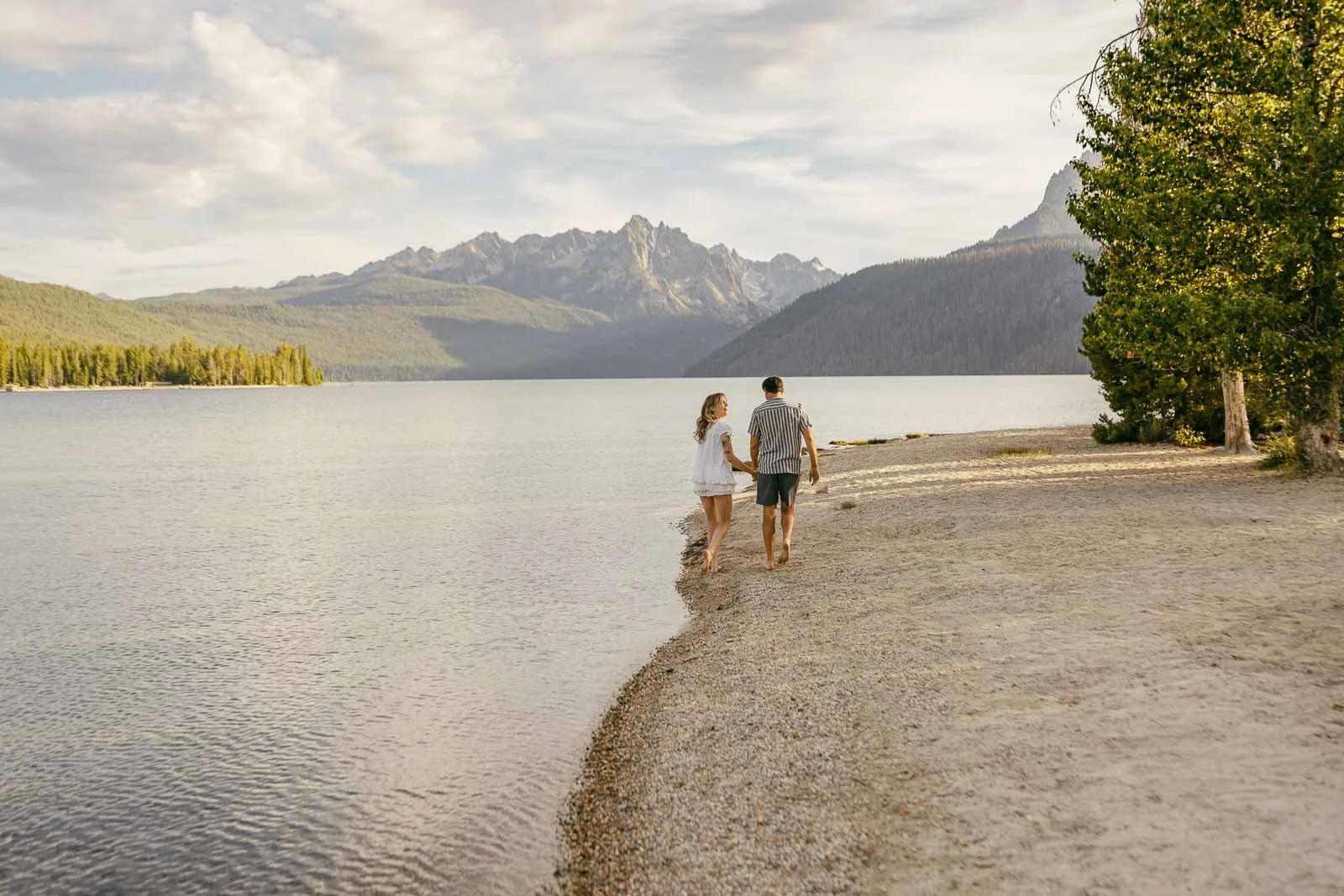  A couple walks hand-in-hand along a sandy lakeshore with mountains and trees in the background on a partly cloudy day. 