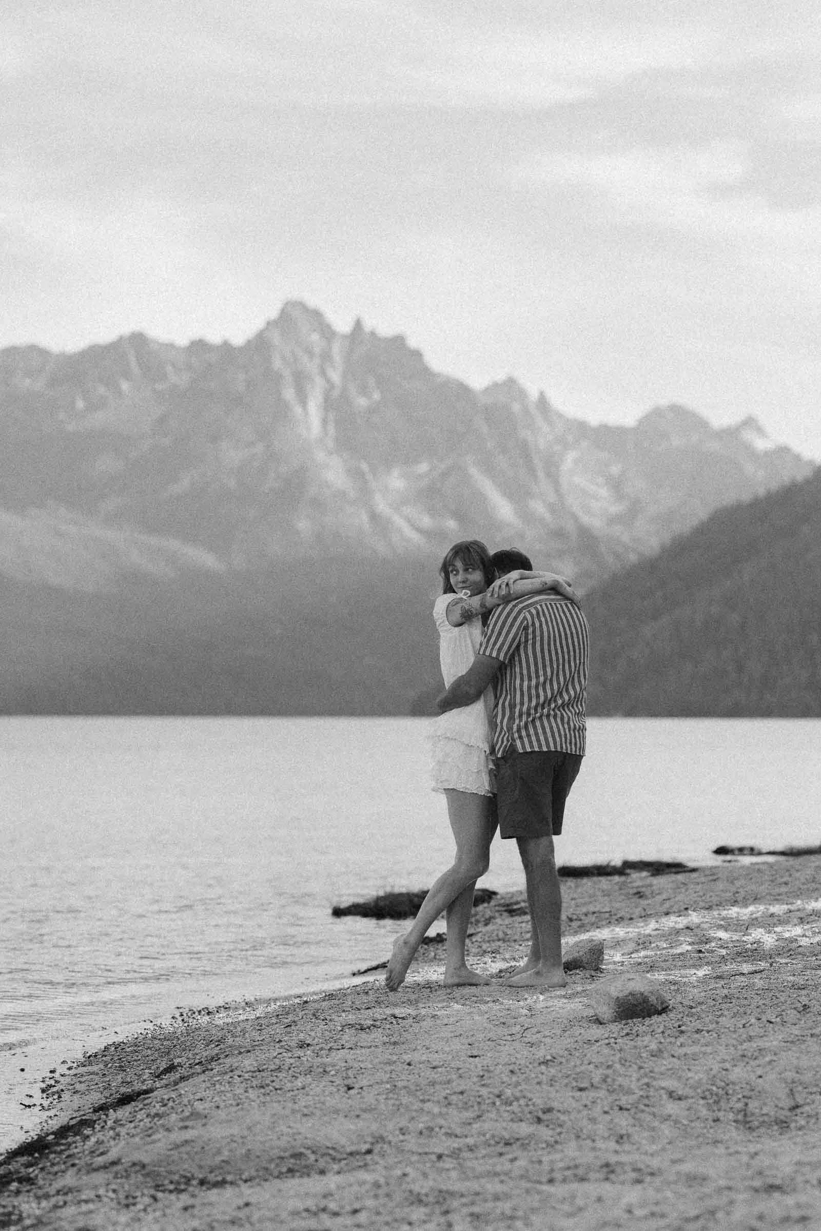  A couple embraces on a lakeshore with mountains in the background. The scene is black and white, and both people are barefoot, standing on the sandy edge of the water. 
