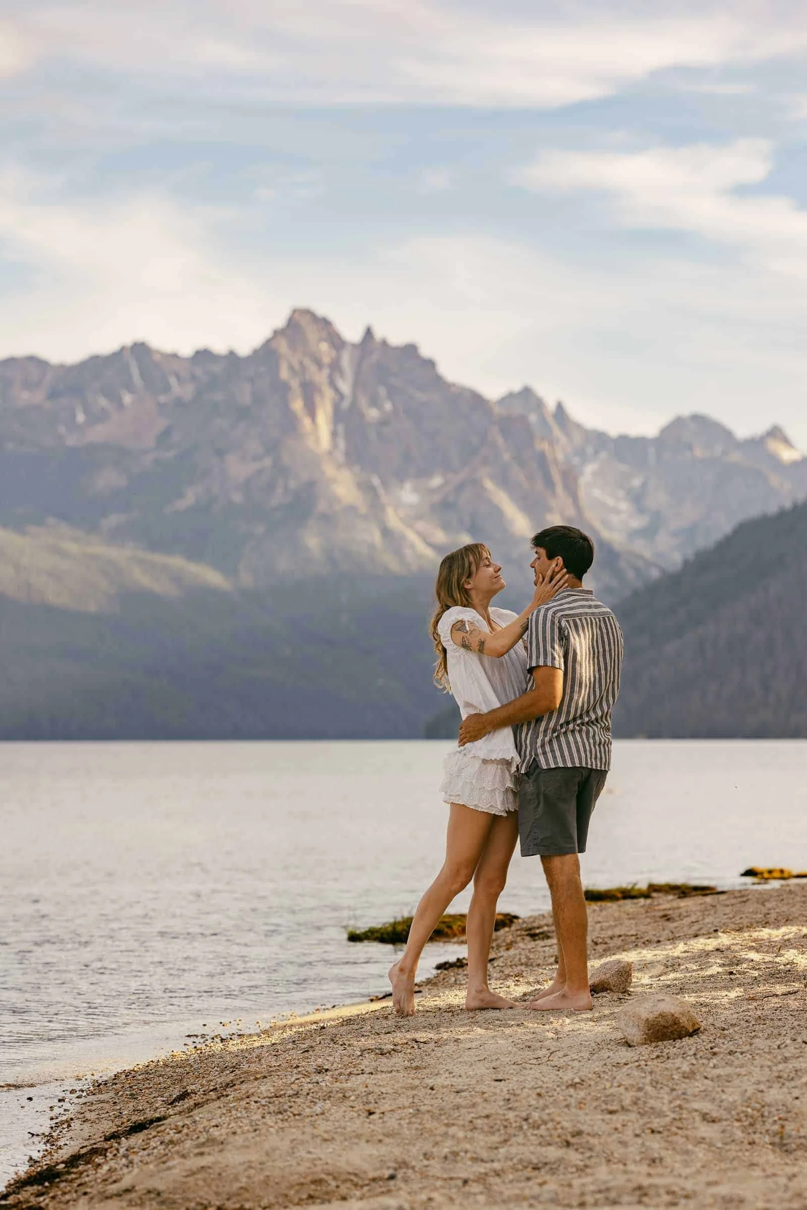  A couple stands barefoot on a lakeshore, embracing and smiling with mountains in the background under a partly cloudy sky. 