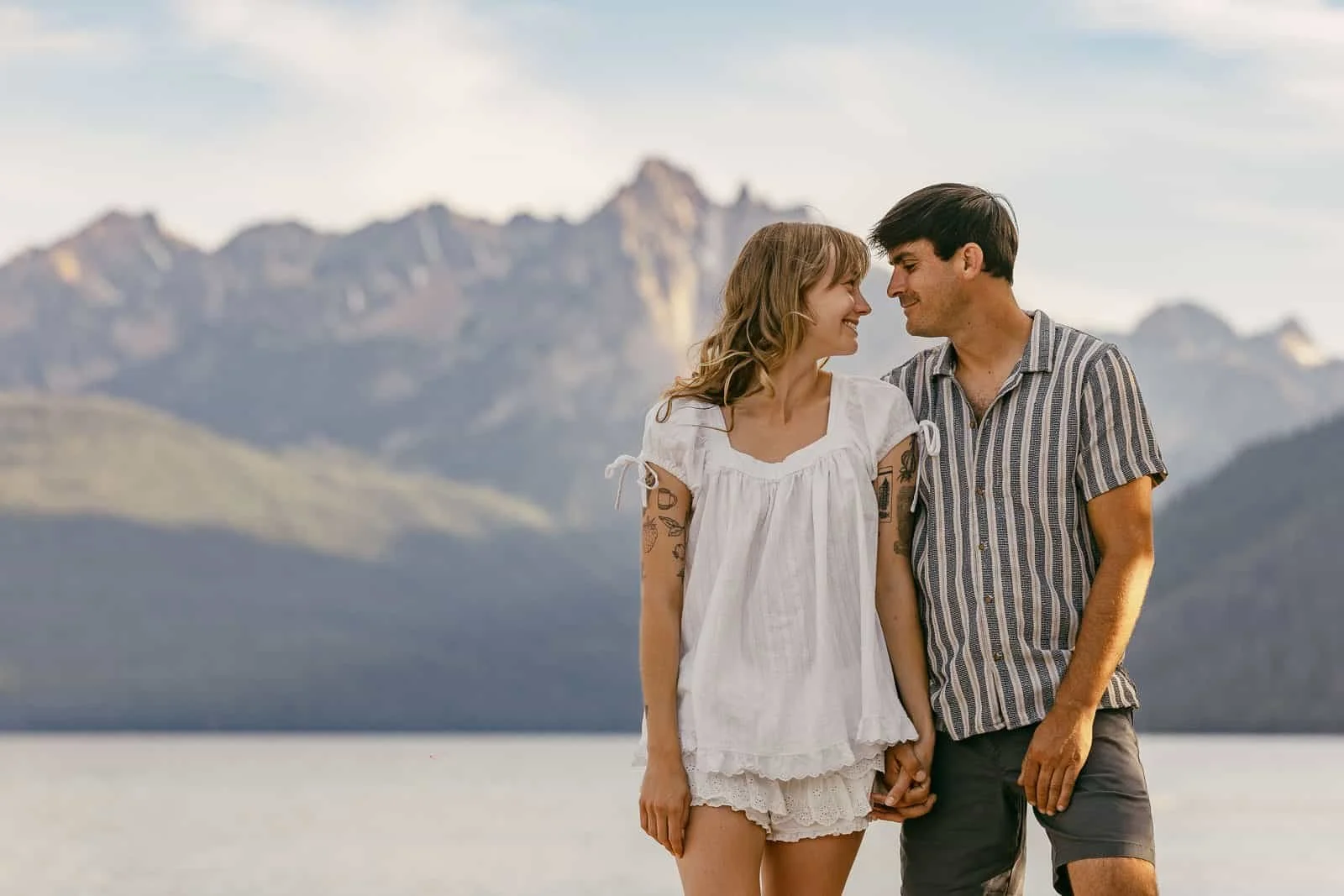  A couple stands hand in hand, smiling at each other, with mountains and a lake in the background under a partly cloudy sky. 