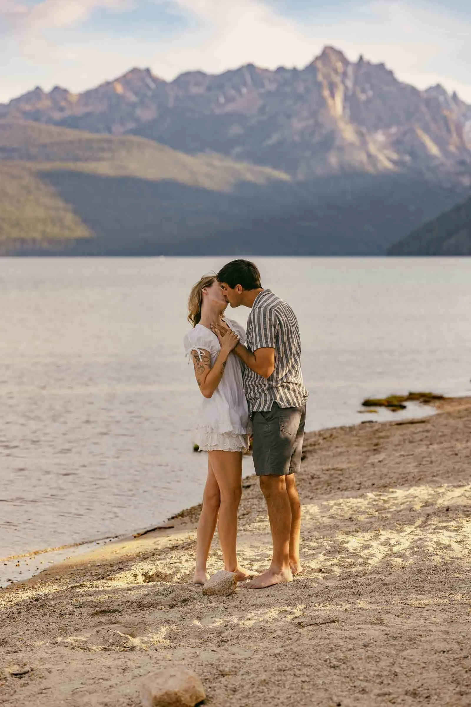  A couple stands barefoot on a sandy lakeshore, holding hands and sharing a kiss, with mountains and a calm lake in the background under a partly cloudy sky. 