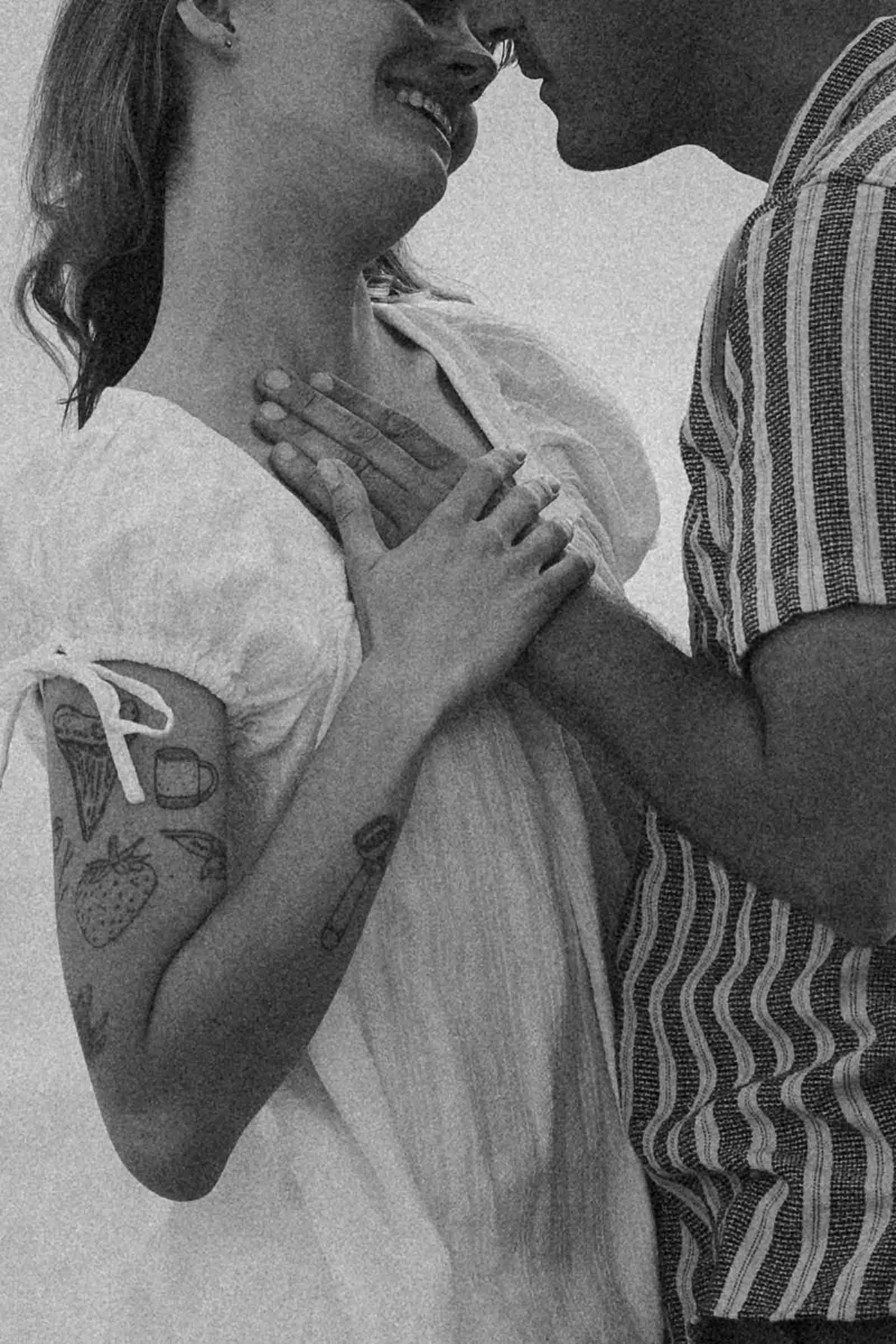  A grayscale photo of a smiling couple standing close, touching faces and hands. The woman wears a light dress with tattoos on her arm; the man wears a striped shirt. 