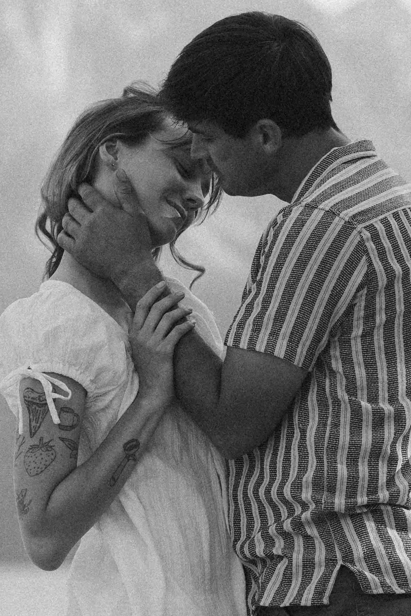  A couple embraces closely, gazing into each other’s eyes with gentle smiles. The woman has visible arm tattoos and wears a light dress; the man wears a striped shirt. The black-and-white photo feels intimate and tender. 