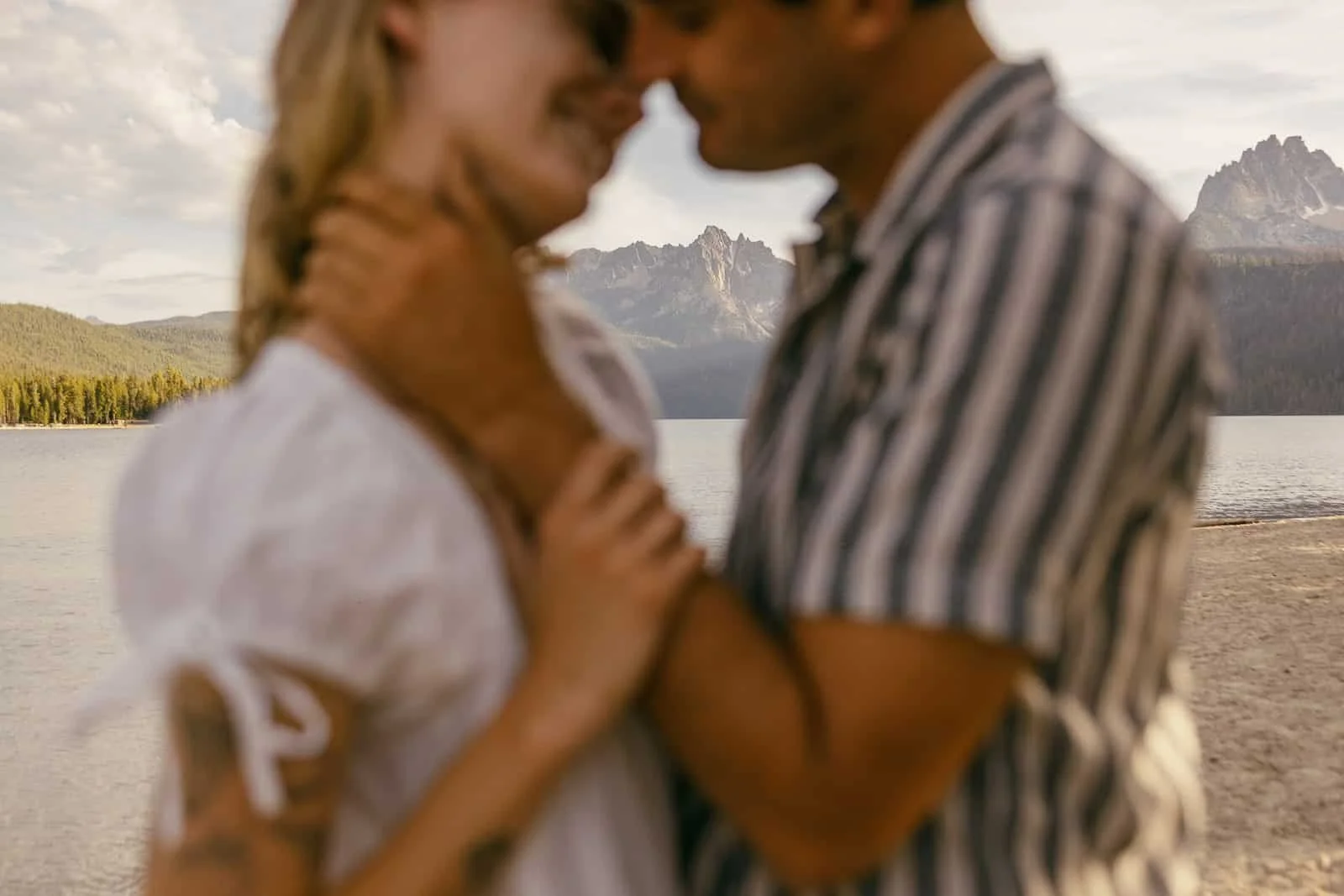  A couple embraces closely and smiles on a lakeshore, with mountains and trees in the blurred background under a cloudy sky. 