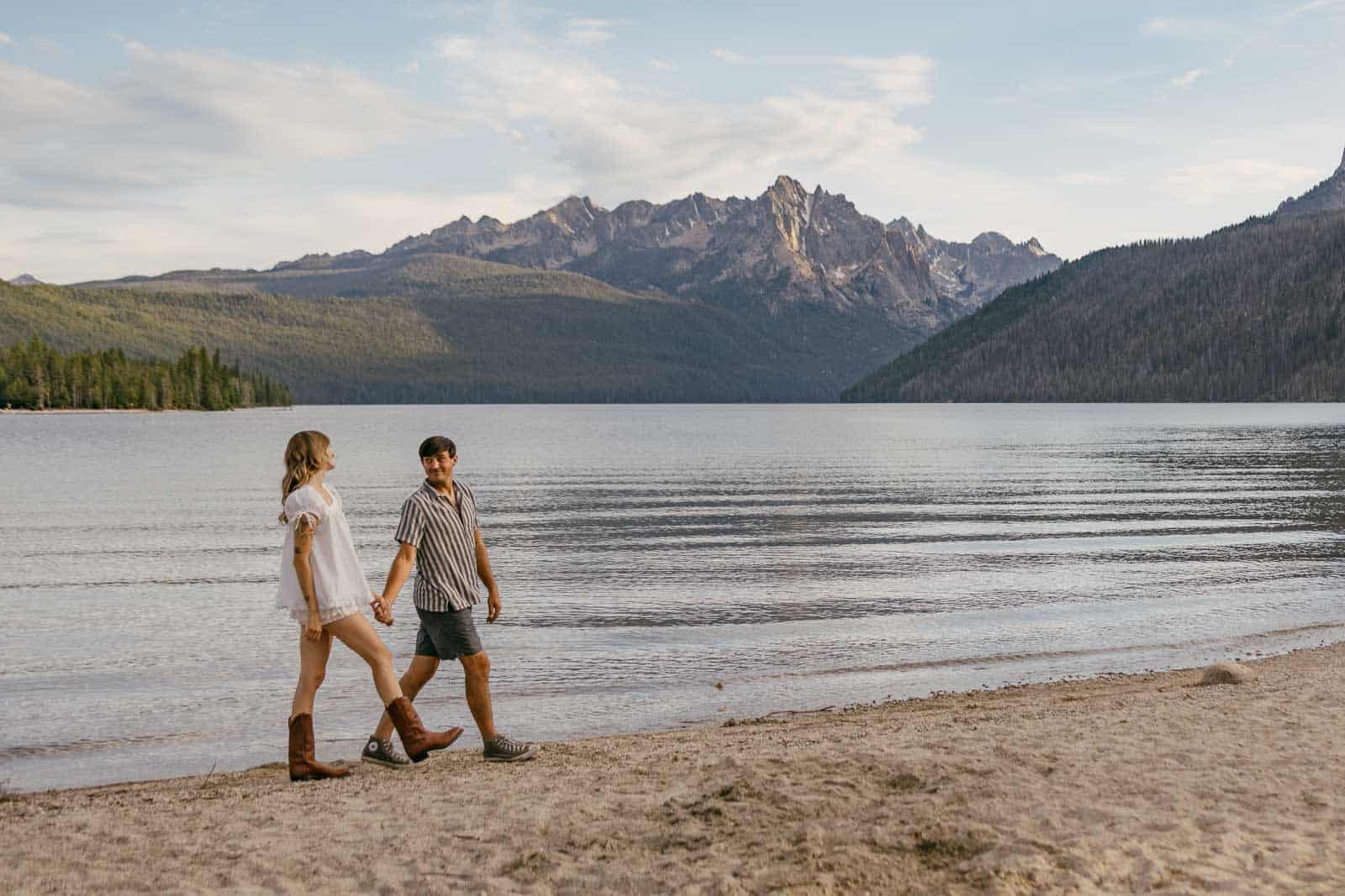  A couple walks hand in hand along a sandy lakeshore with calm water and forested mountains in the background under a blue sky. 