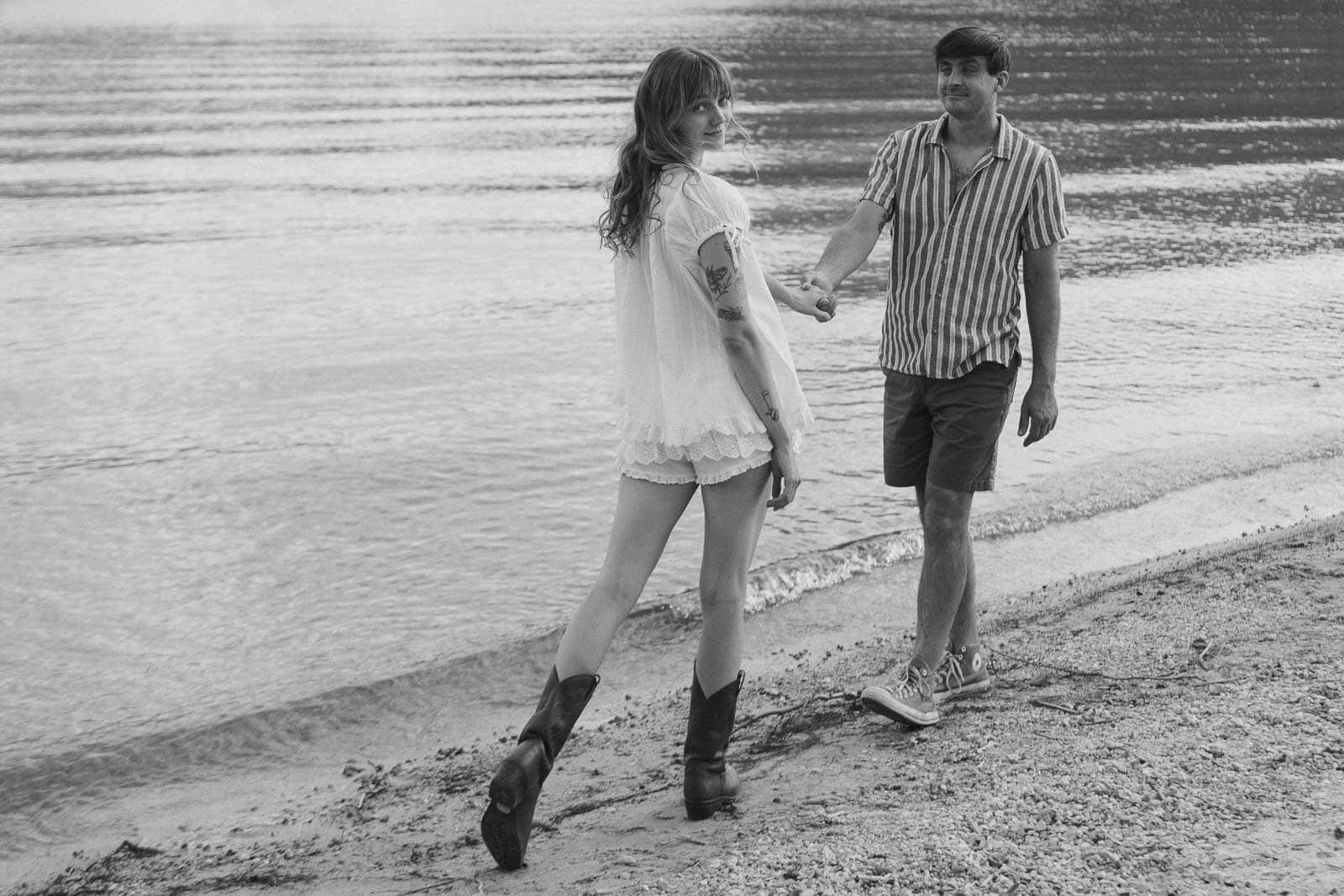  A woman in boots and a light outfit holds hands with a man in a striped shirt and shorts as they walk along a calm lakeshore. The image is in black and white. 