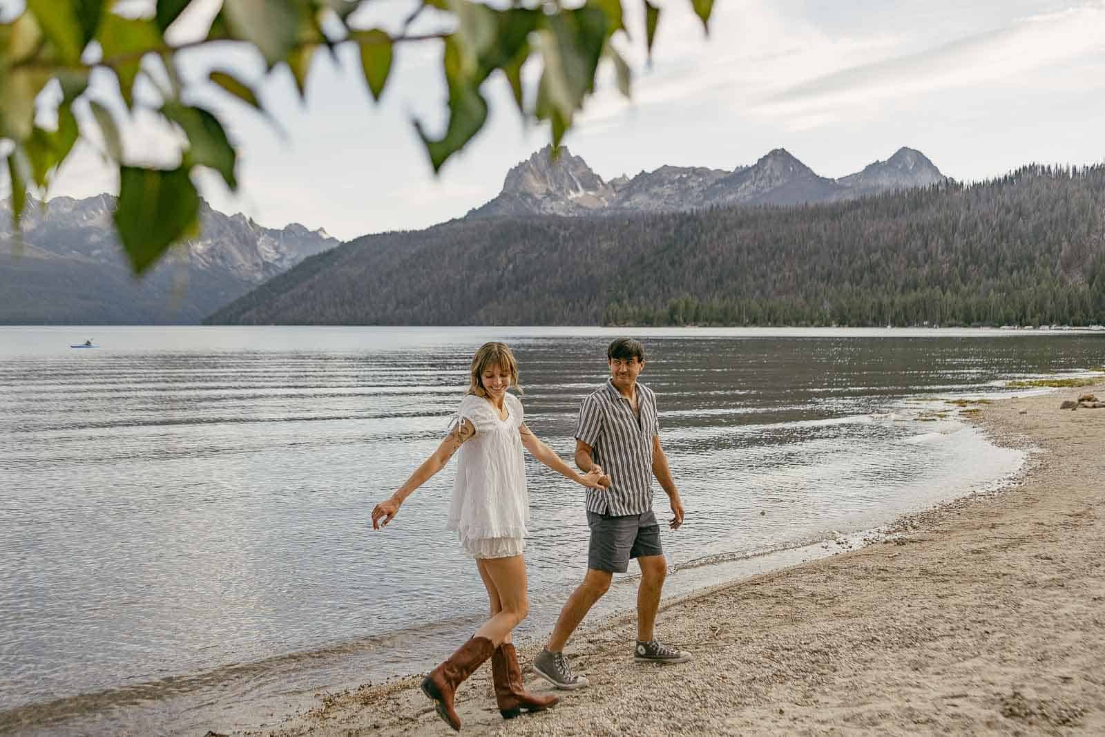  A young couple holding hands walks along a sandy lakeshore with mountains and forests in the background. A few green leaves hang at the top left corner of the image. 