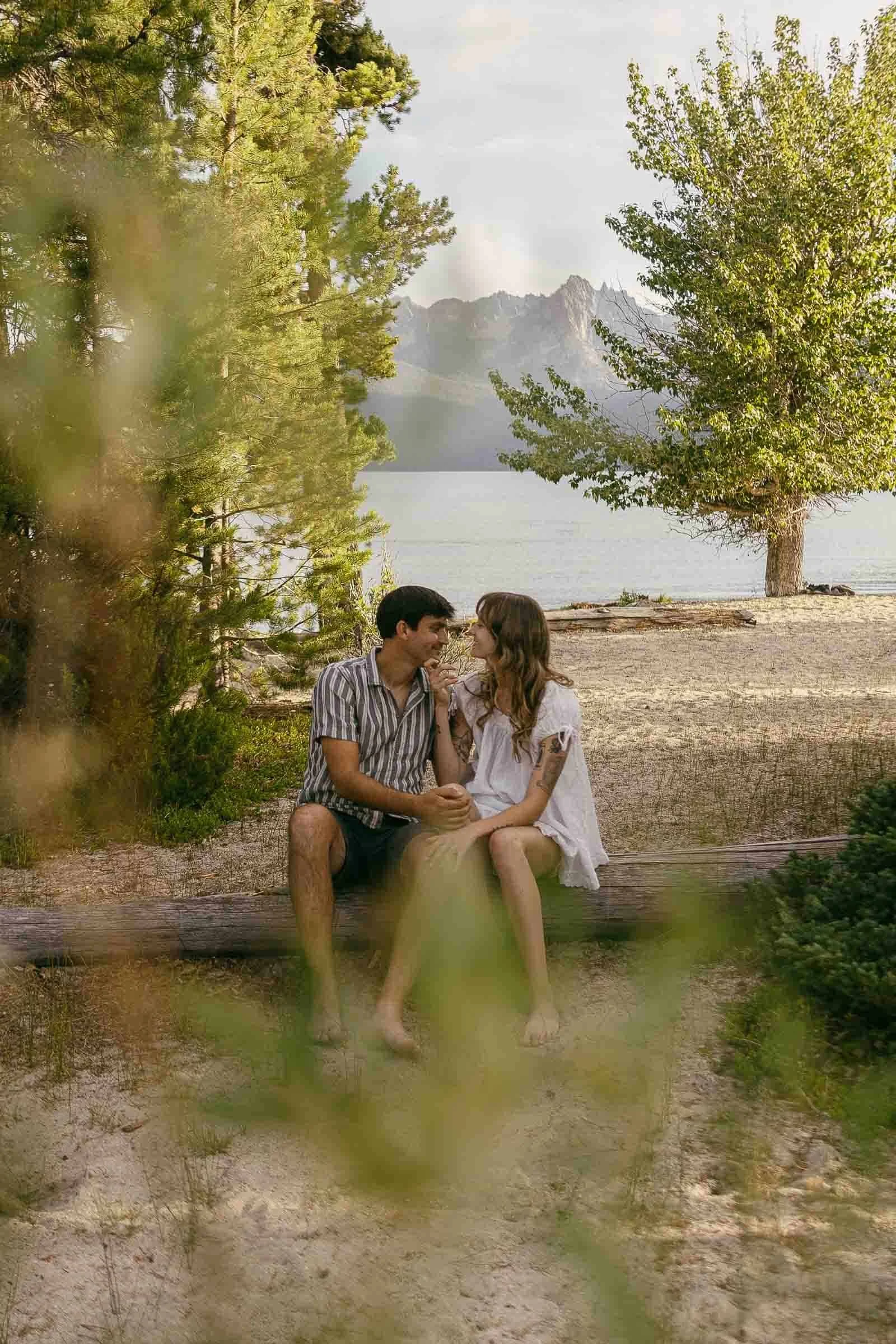  A couple sits on a log at a lakeshore, holding hands and looking at each other, surrounded by trees and mountains in the background. 