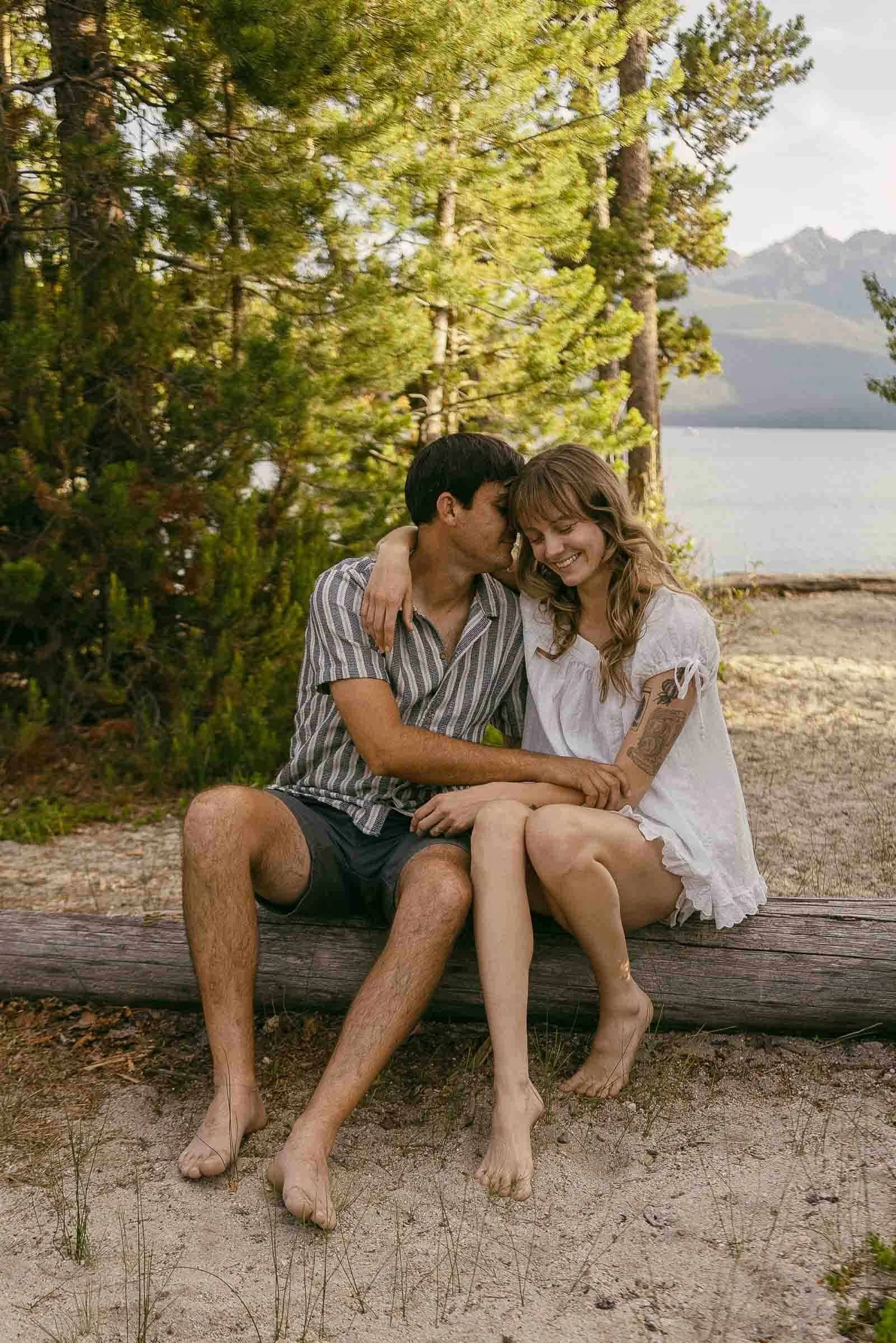  A barefoot couple sits on a log by a lake, surrounded by trees. The man kisses the woman’s cheek while she smiles, both looking relaxed and happy in casual summer clothes. 