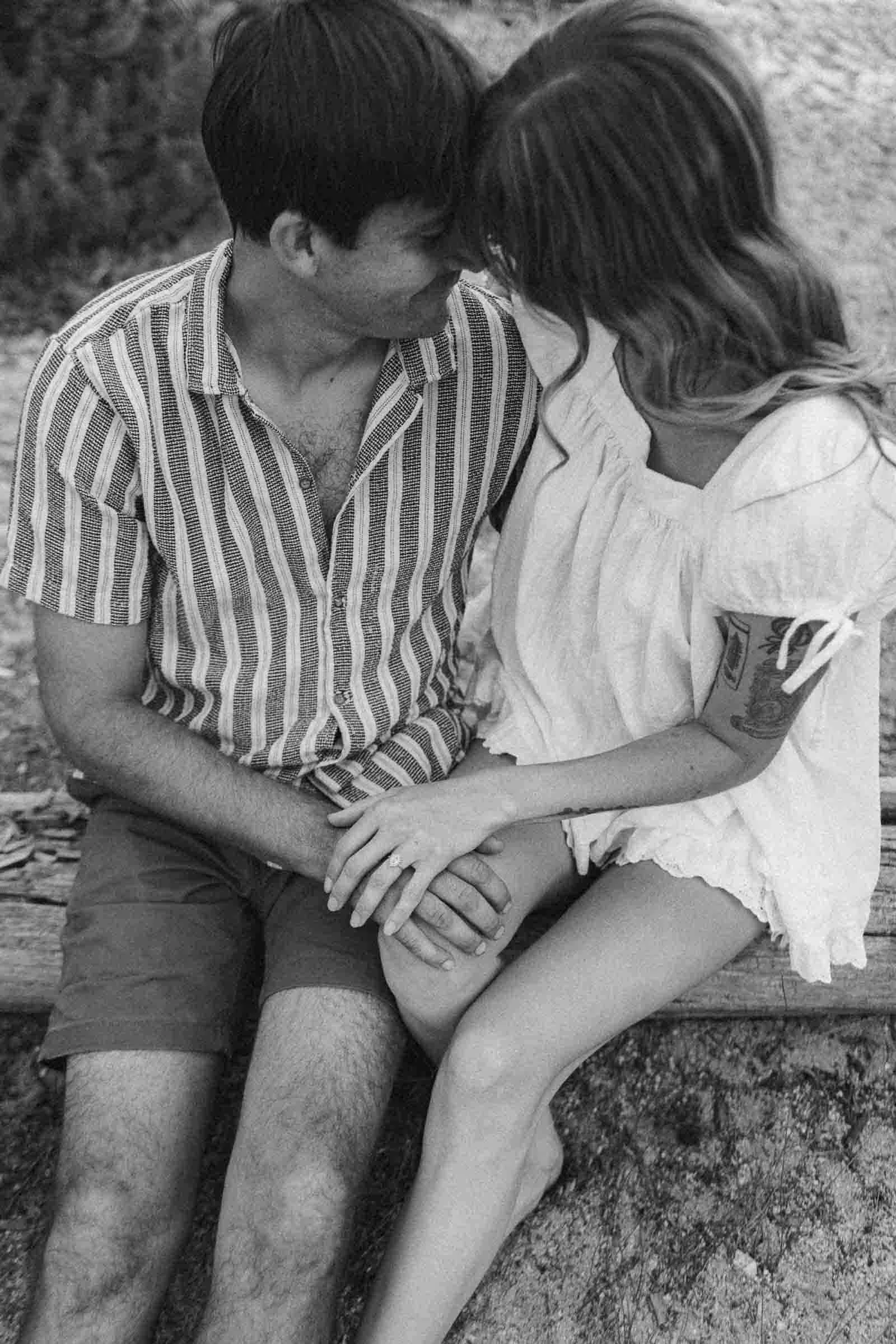  A couple sits closely on a bench, holding hands and touching foreheads, sharing an intimate moment. The photo is black and white. 