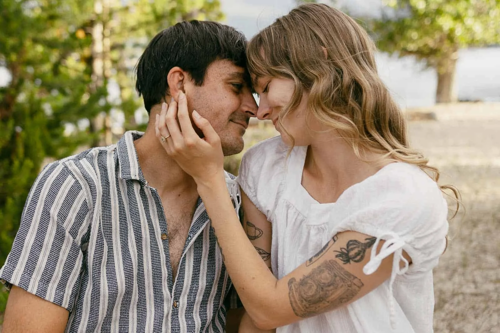 A couple sits close together outdoors, touching foreheads and smiling softly. The woman gently holds the man’s face, displaying a ring on her finger and tattoos on her arm. Trees and water are visible in the background. 