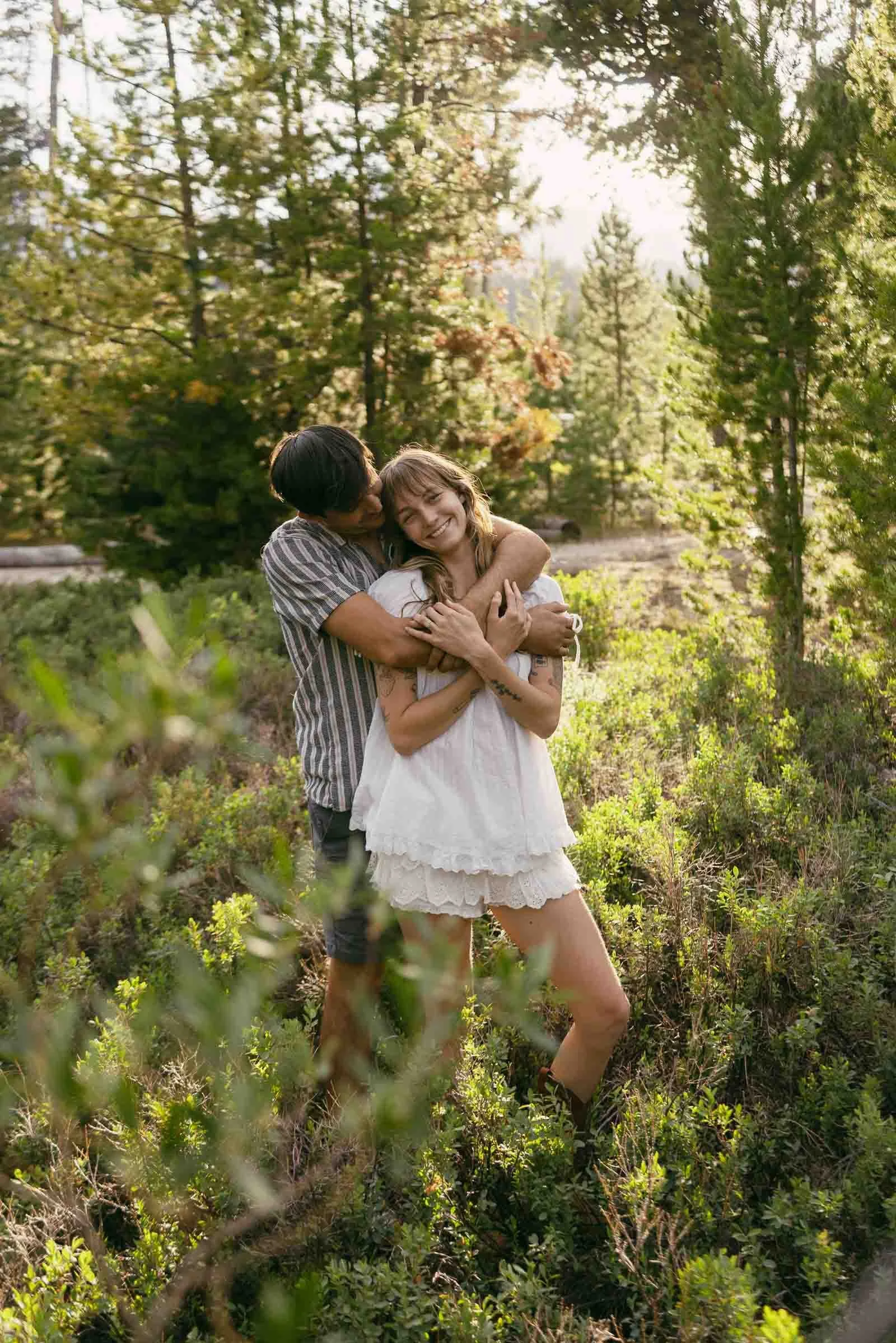  A couple stands in a sunlit forest. The man hugs the smiling woman from behind as she stands in front, wearing a white dress. Lush green trees and plants surround them. 