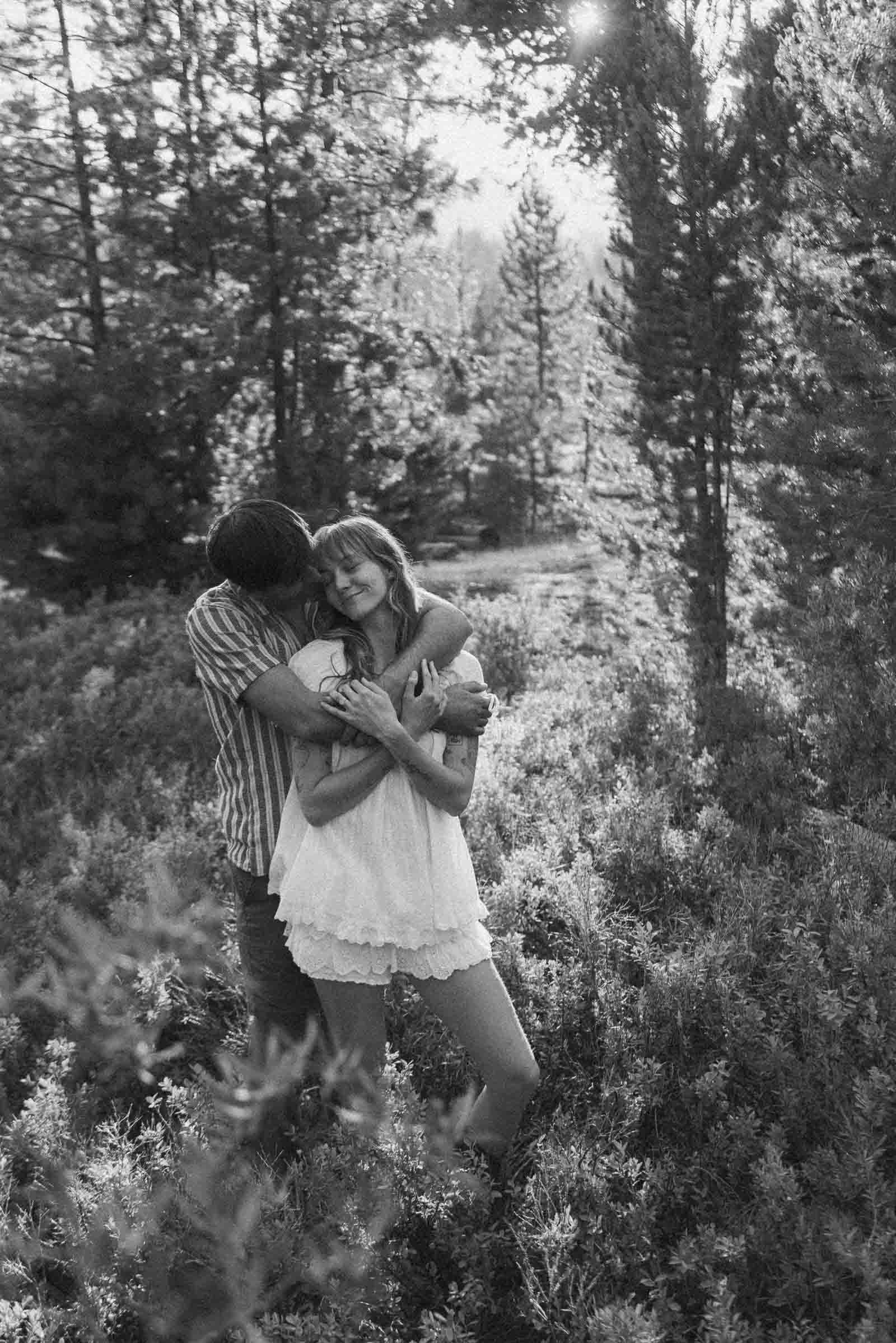  A couple stands in a sunlit forest, embracing and smiling. The woman wears a white dress, while the man hugs her from behind. Trees and bushes surround them in this black-and-white photo. 