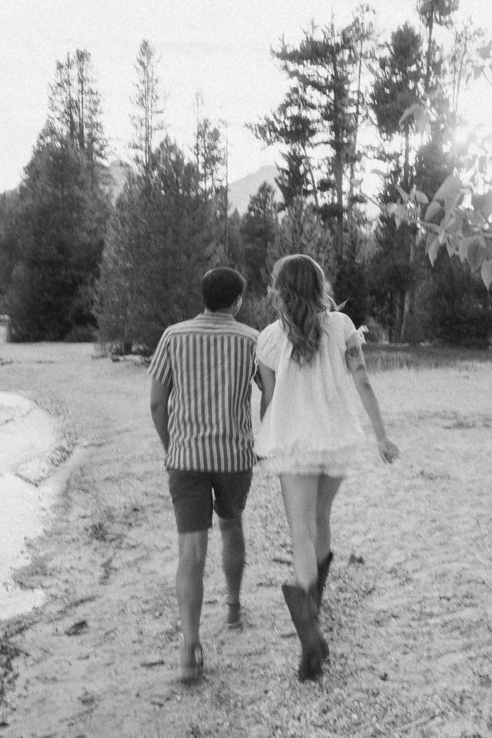  A man and woman walk side by side on a sandy lakeshore, surrounded by trees. The woman wears a short dress and boots; the man wears a striped shirt and shorts. The image is in black and white. 