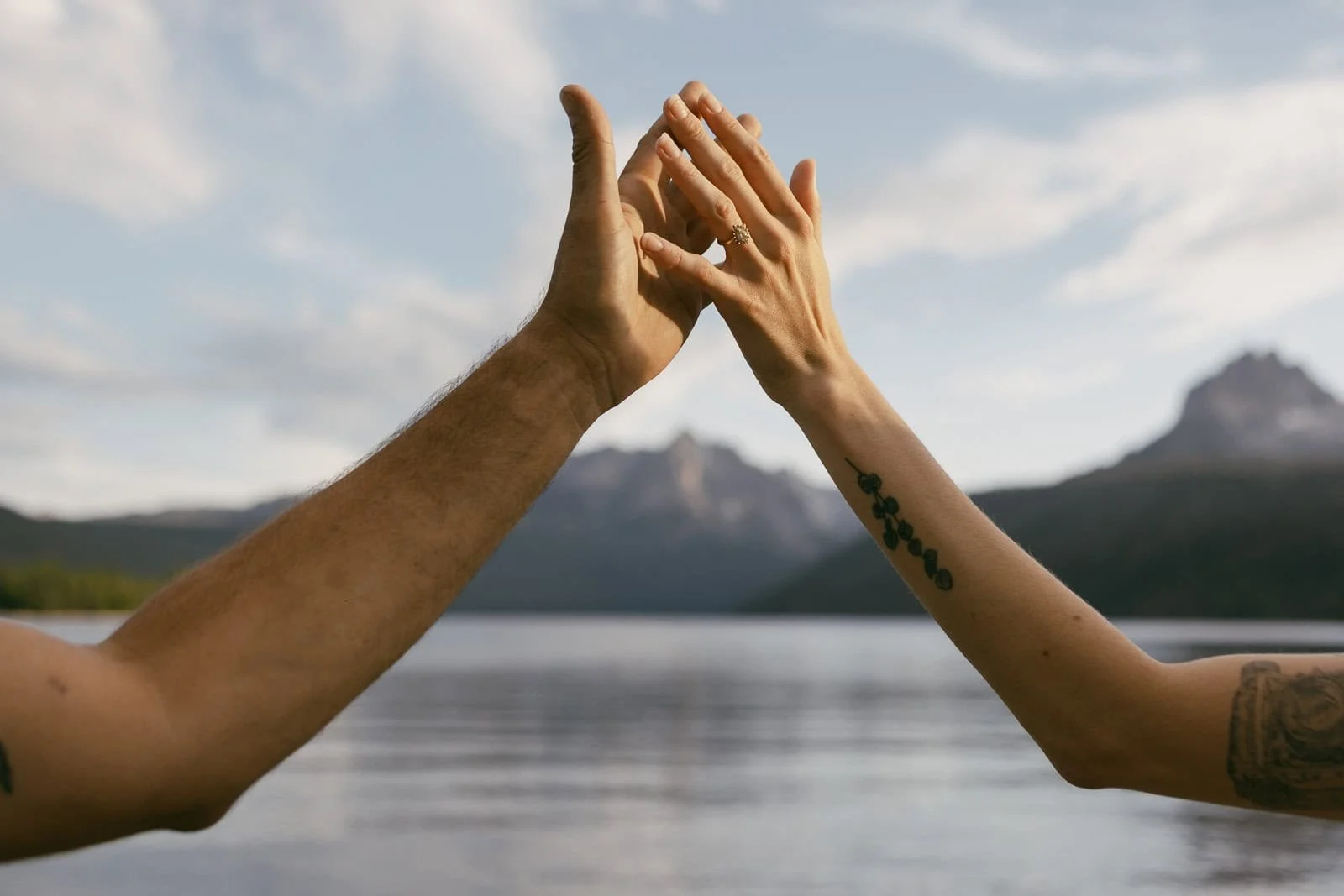  Two people’s hands touching palm to palm in front of a lake with mountains and a cloudy sky in the background. One arm has a small plant tattoo. 
