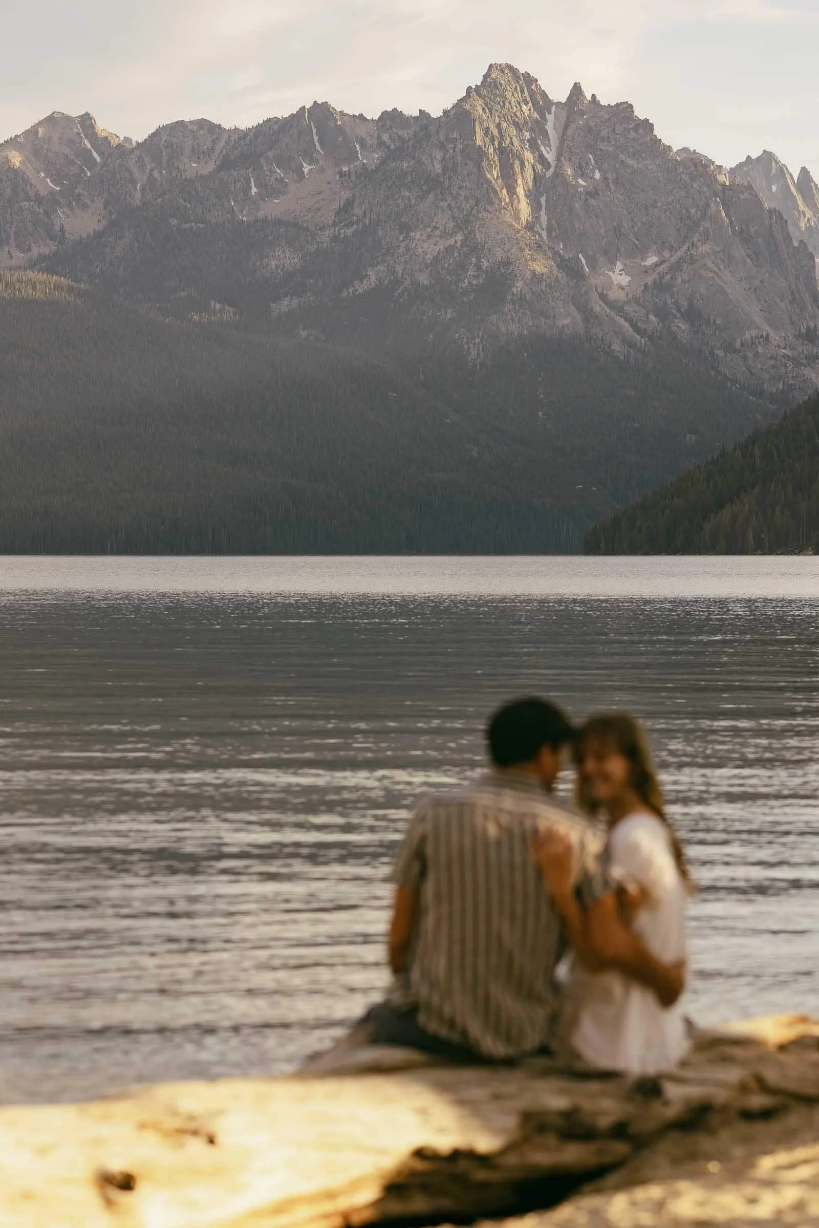  A couple sits on a log by a calm lake, facing each other with mountains and trees in the background. The couple is out of focus, while the mountains and lake are sharp and clear. 