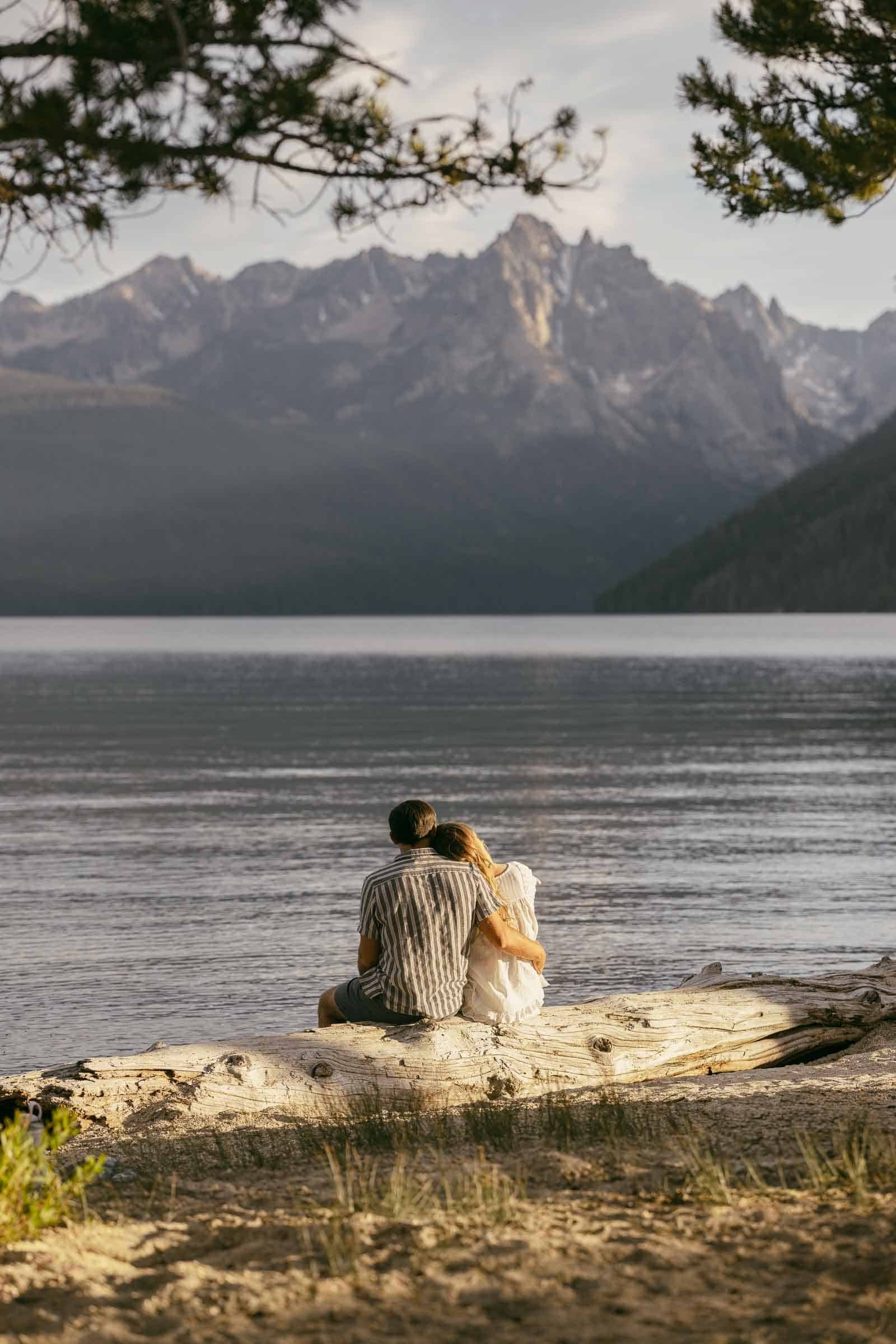  A couple sits on a log by a calm lake, facing distant mountains under a cloudy sky, framed by pine tree branches. 