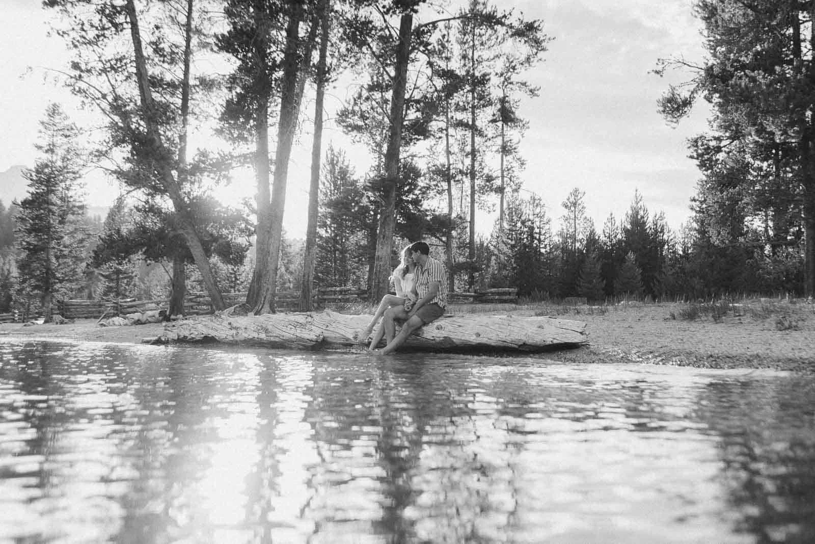  A couple sits on a log by a lakeshore, feet in the water, surrounded by tall trees and nature. The scene is peaceful and sunlit, in black and white. 
