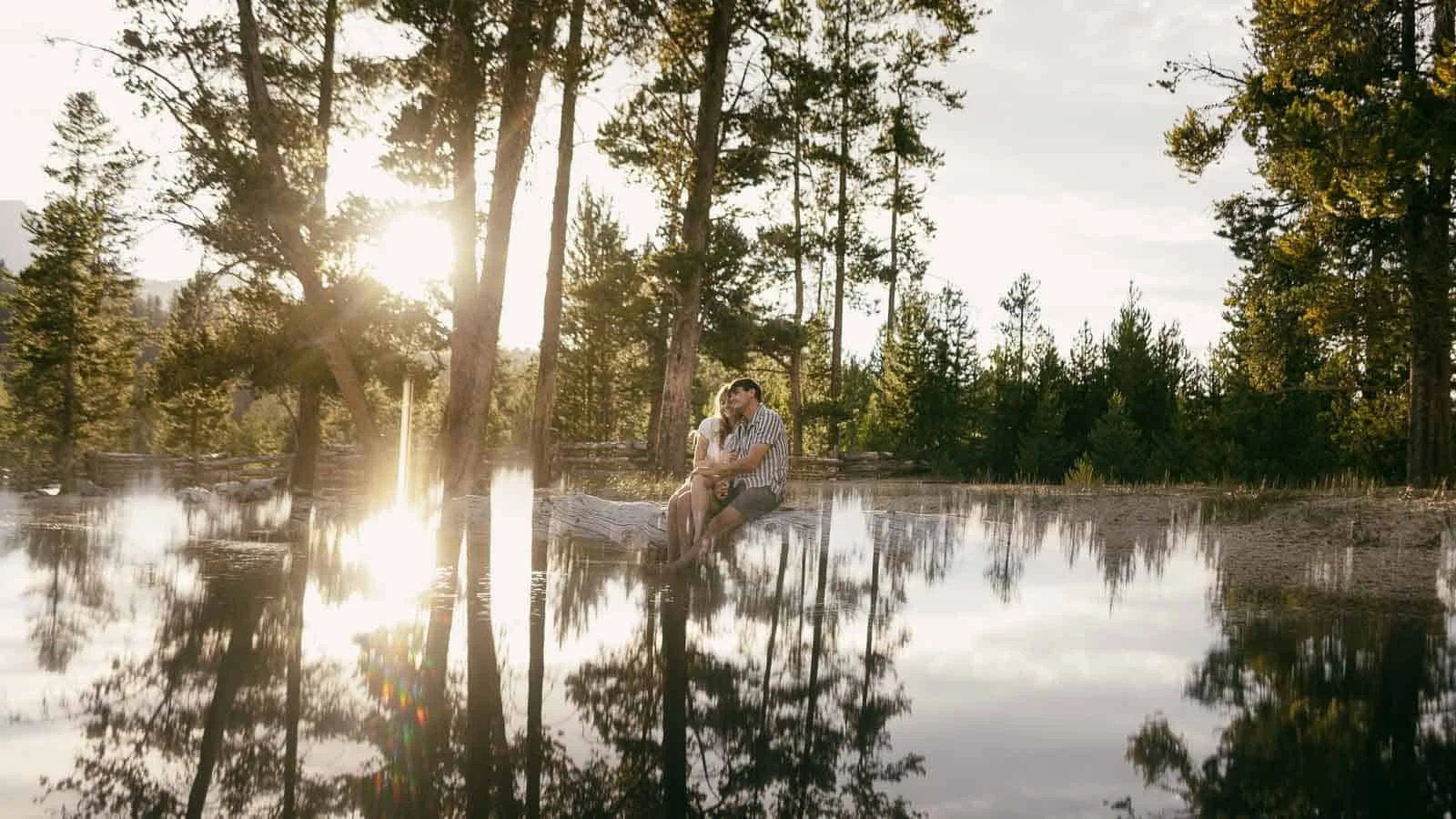 A couple sits close on a fallen log stretched over a calm, tree-lined lake, holding each other as the setting sun glows on the water behind them.