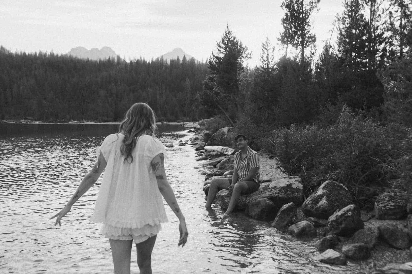  A person in a white dress stands in shallow water, facing another person sitting on rocks by a lakeshore, surrounded by trees and distant mountains, in black and white. 