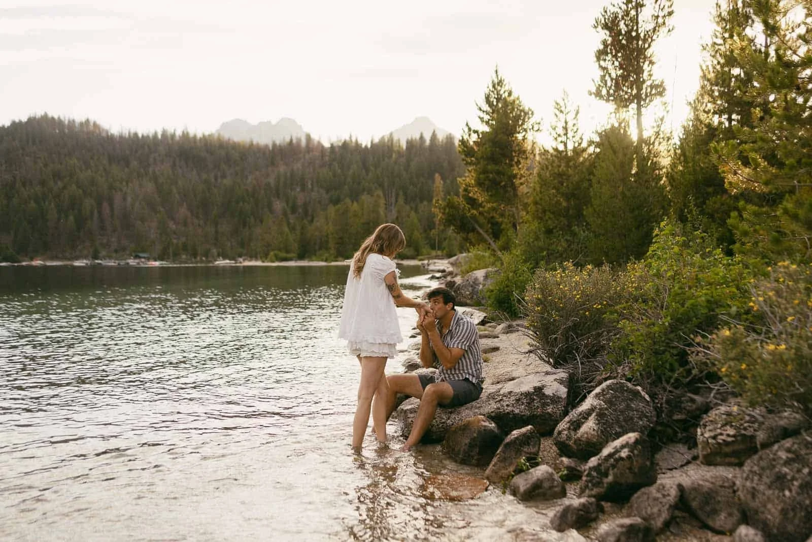  A woman in a white dress stands in shallow lake water while a man sits on a rock, playing guitar. They are surrounded by trees and mountains under a cloudy sky. 