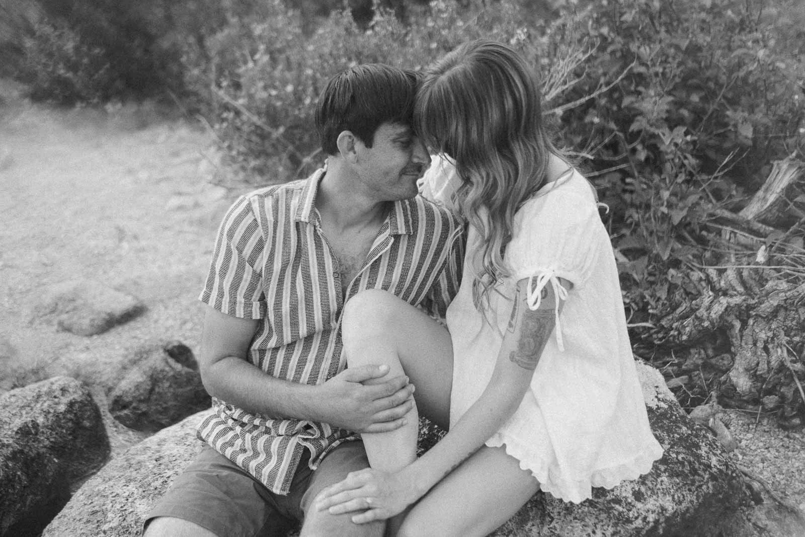  A couple sits close together on rocks outdoors, touching foreheads and smiling, surrounded by greenery. The photo is in black and white. 
