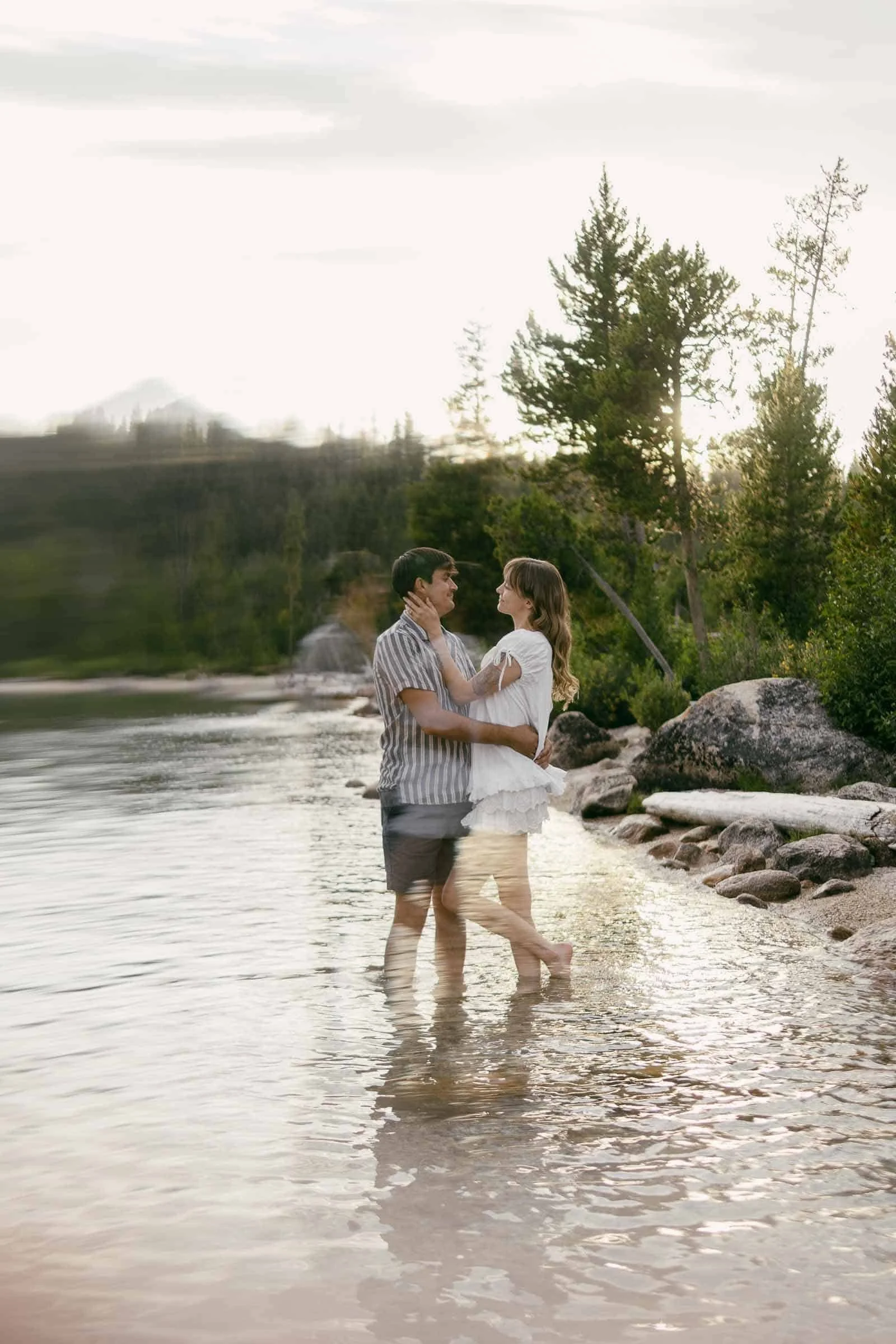  A couple stands in shallow lake water, smiling at each other and embracing. Trees and rocks line the shore, and the scene has a soft, dreamy blur effect. 