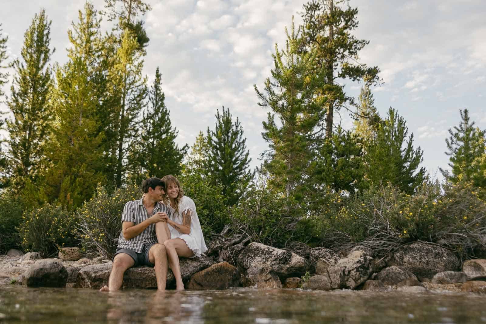  A couple sits on rocks by a lakeshore, surrounded by tall pine trees and greenery, sharing a cozy moment under a partly cloudy sky. 