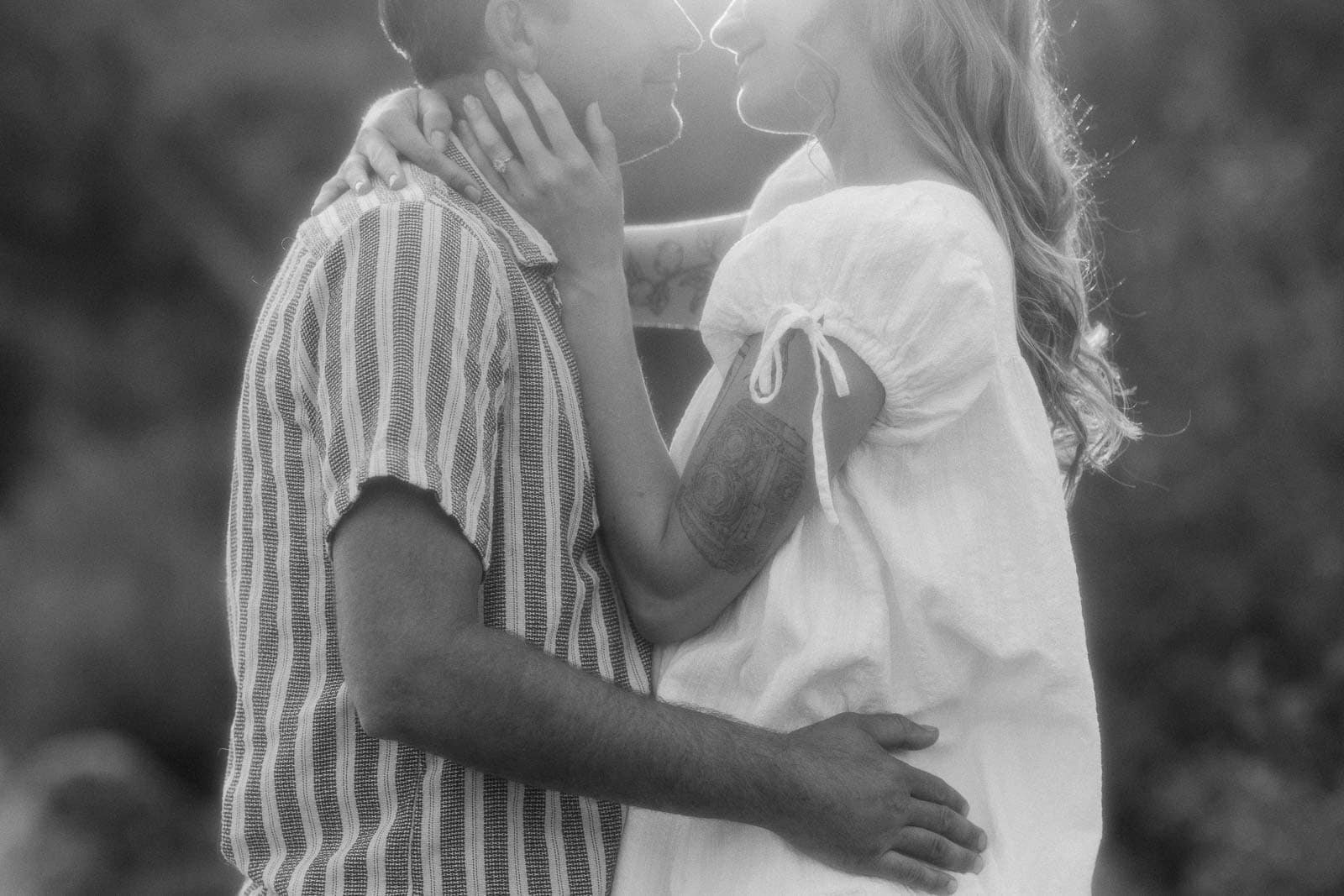  A black-and-white photo of a couple standing close, smiling and touching each other’s faces affectionately, with soft light and a blurred outdoor background. 